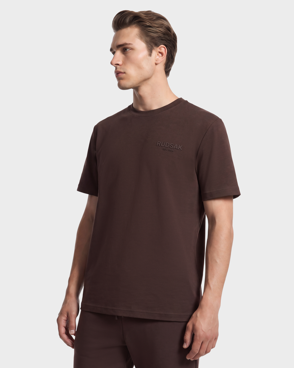 A man wearing a RUDSAK GRANT - 6525036 Espresso breathable cotton, short-sleeved dark brown crewneck tee with a small logo on the chest.
