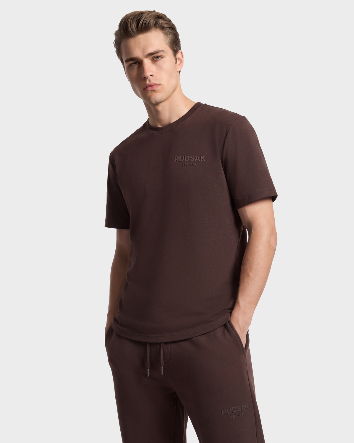 A man wearing the GRANT - 6525036 Espresso breathable cotton crewneck tee and matching drawstring sweatpants, both featuring an embossed logo on the front.