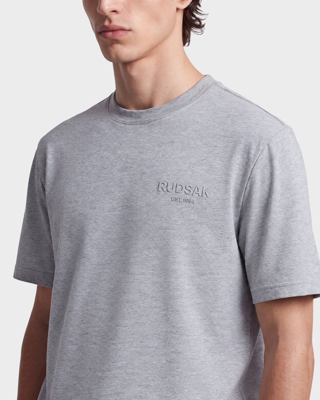 A person wearing the Rudsak GRANT - 6525036 Heather Grey men’s tee, featuring short sleeves and a classic crewneck design, crafted from premium cotton with “RUDSAK EST. 1994” embroidered on the chest. The background is plain white.