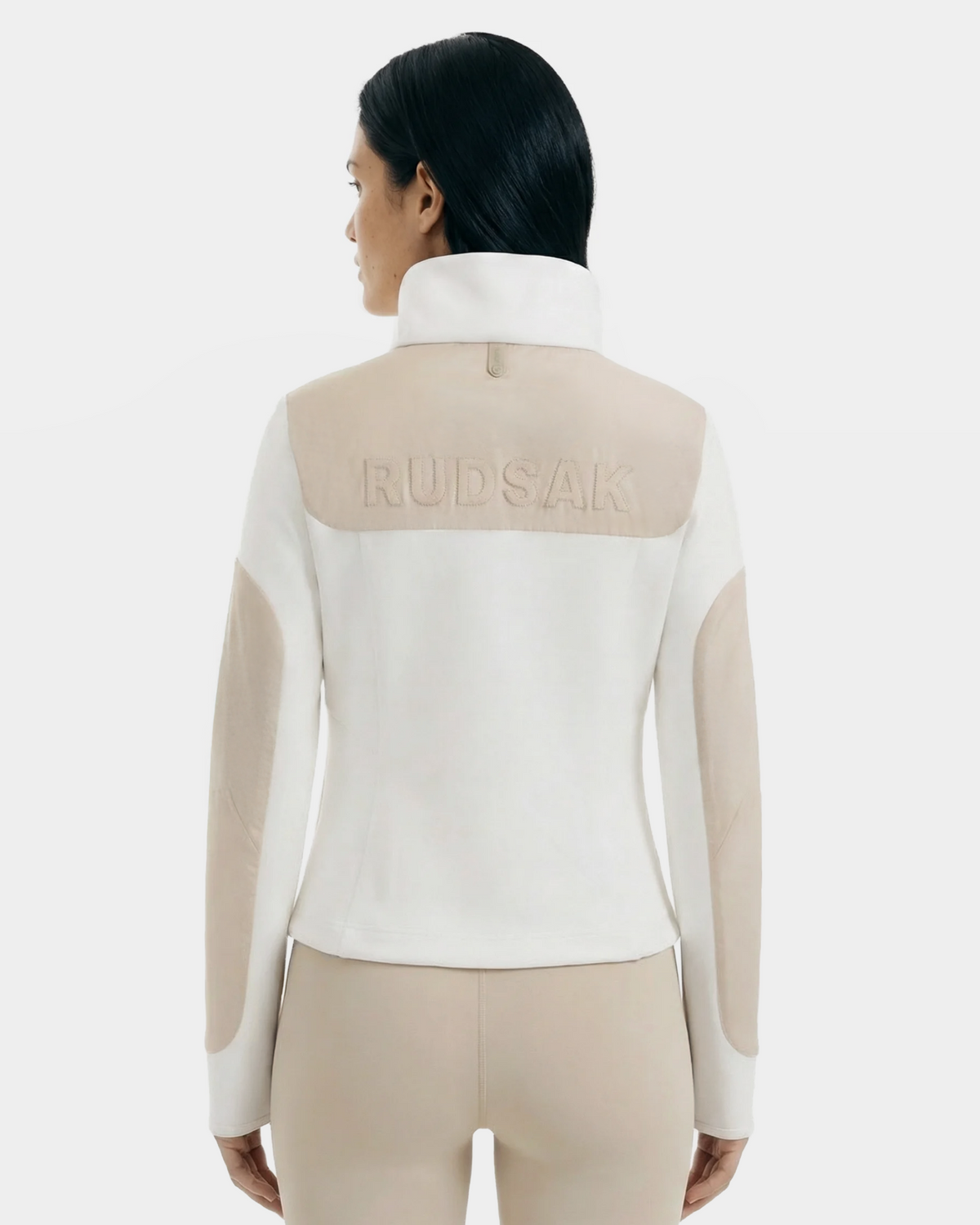 A woman with straight black hair wears the Rudsak GRANADA - 8126023 Vanilla sweater jacket in white and beige, embroidered with RUDSAK on the back, paired with beige pants. She stands facing away against a plain light background.