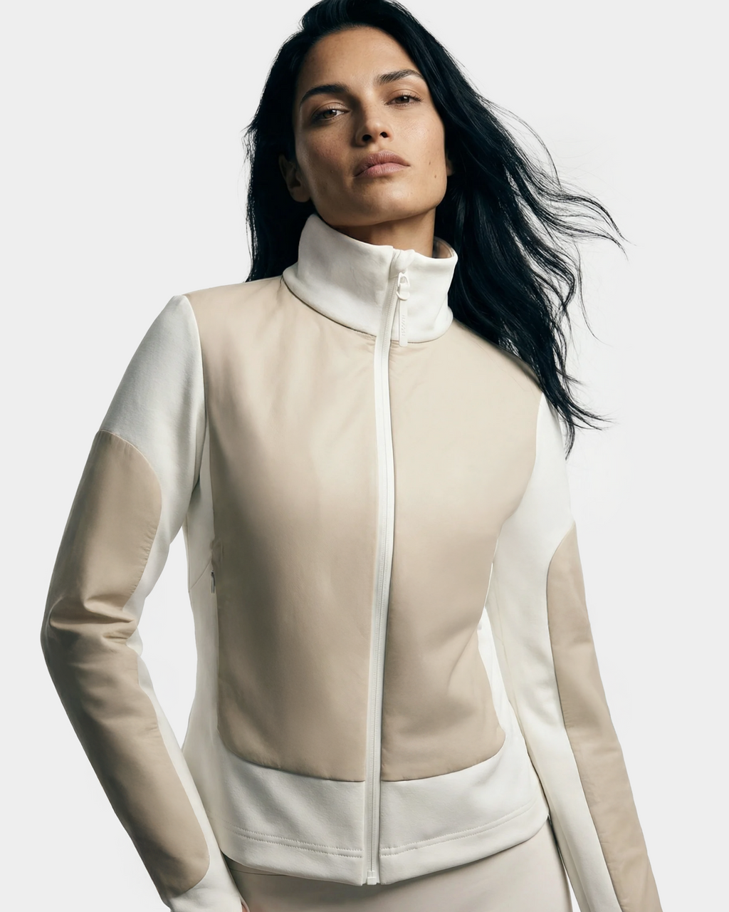 A woman with long dark hair models the Rudsak GRANADA - 8126023 Vanilla: a fitted, high-neck lightweight jacket in beige and white color blocks with mixed-media construction, set against a plain light background.