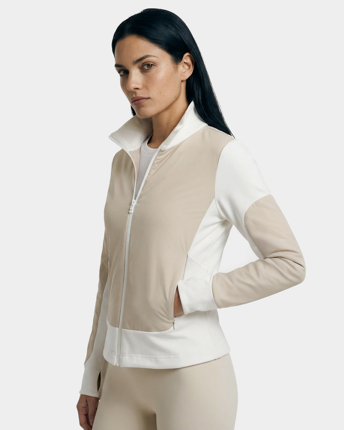 A woman with long dark hair wears the Rudsak GRANADA - 8126023 Vanilla zip-up sweater jacket and matching leggings, standing with one hand in her pocket against a plain light background, exuding sporty-chic everyday ease.