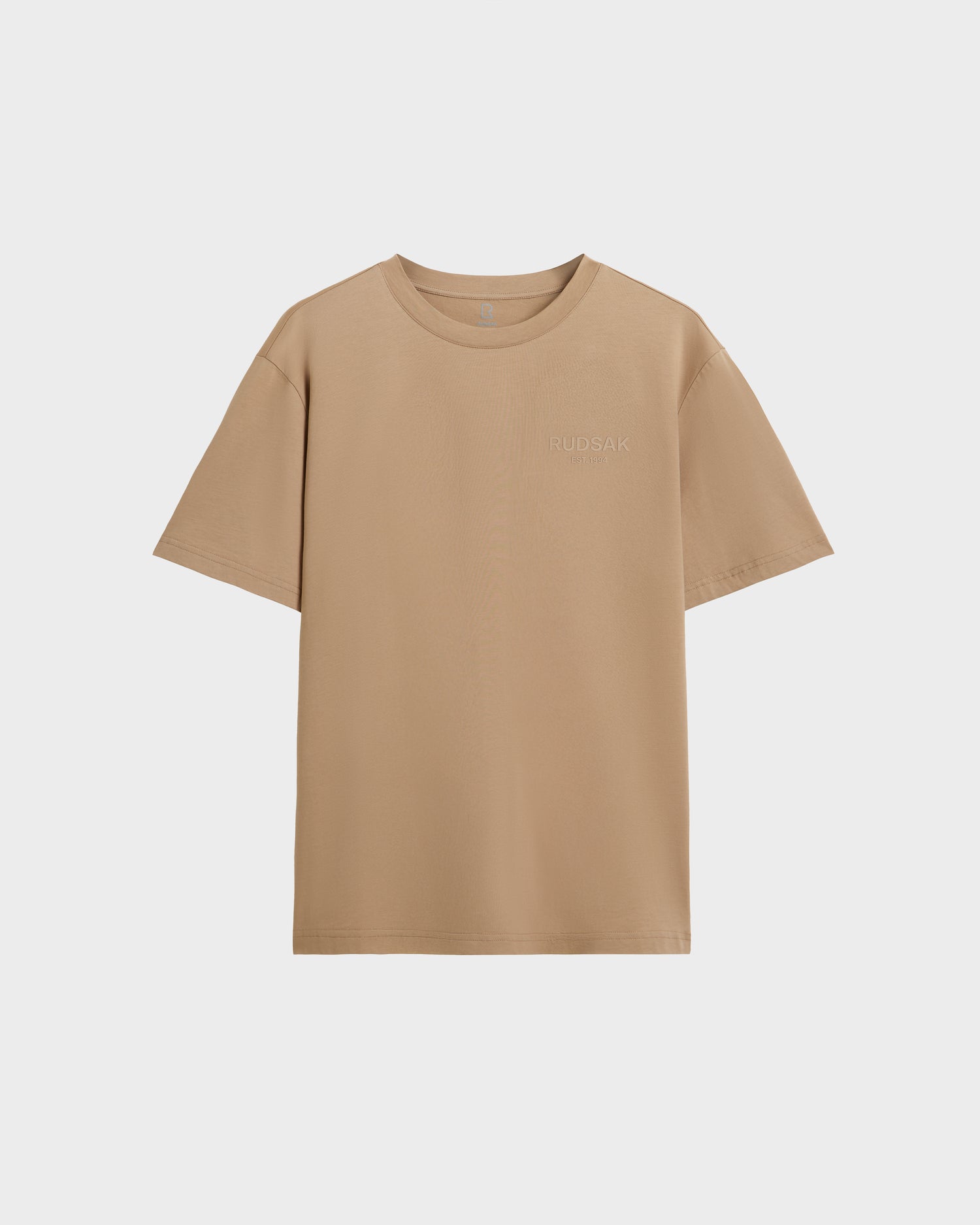 The Rudsak GRANT - 6525036 Almond is a premium cotton men’s beige crewneck tee with short sleeves and minimal embroidered text on the left chest, shown on a white background.