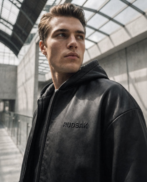 A man wearing a black leather hooded jacket with 'RUDSAK' debossed on the chest, standing in a modern concrete walkway under a glass roof.