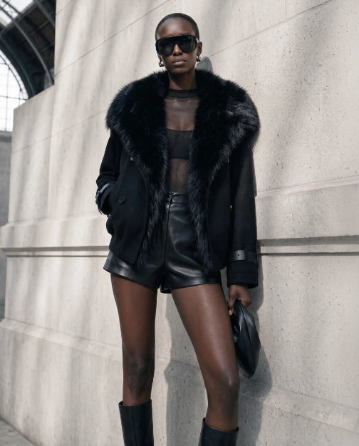 A model wearing a black faux-fur trimmed jacket, black leather shorts, and black knee-high boots, posing against a light gray concrete wall.