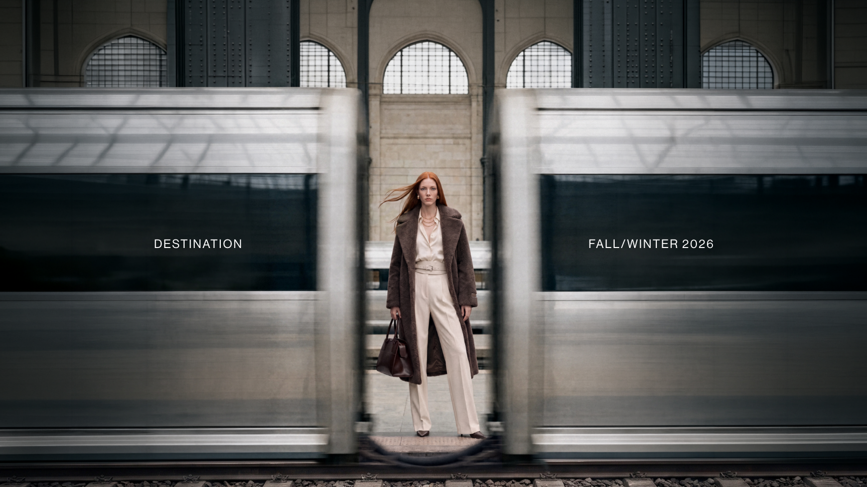 A woman stands on a train platform between two large silver train doors, wearing a long brown coat, cream pants, and carrying a dark handbag. The scene is framed symmetrically with the trains and arched windows behind her.