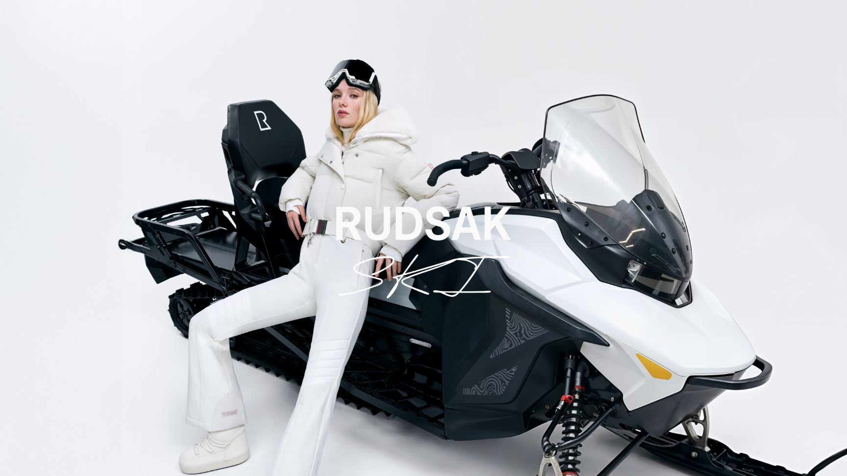 Person in white snow gear sitting on a snowmobile with 'RUDSAK' branding.