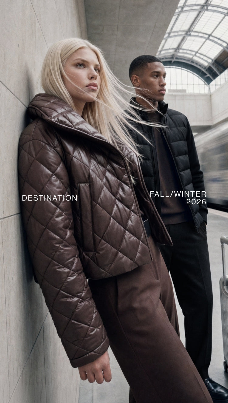 A fashion editorial shot featuring a blonde woman in a brown quilted puffer jacket leaning against a concrete wall, with a man in a black quilted puffer jacket standing behind her in a modern transit station. Text overlays read 'DESTINATION' and 'FALL/WINTER 2026'.