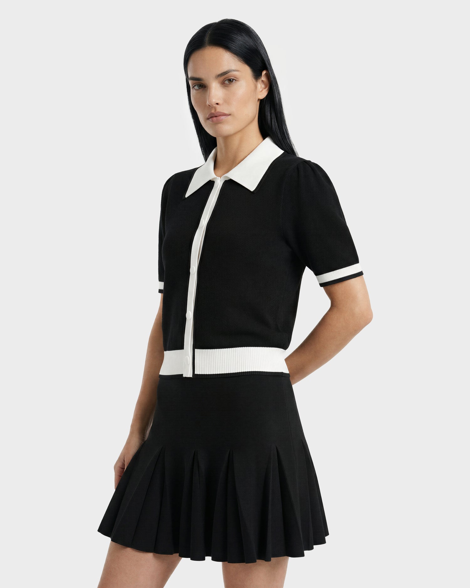 A woman with long black hair models the Rudsak IRIS - 8526012 Black short-sleeve sweater with a contrast collar and buttons, paired with a black pleated skirt, standing against a plain light gray background.
