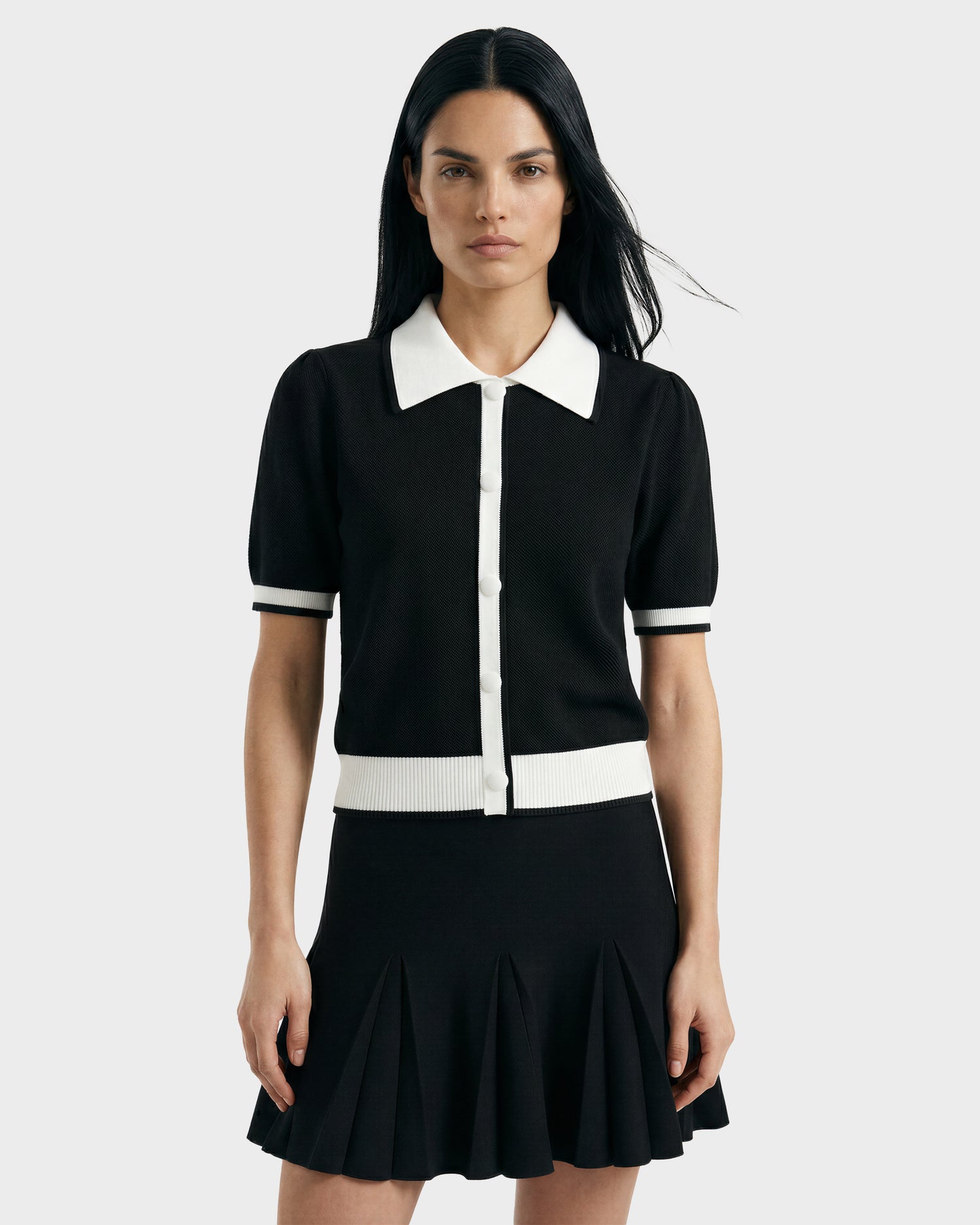A woman with long dark hair wears the Rudsak IRIS - 8526012 Black knit short-sleeve sweater with white trim and contrast collar, paired with a black pleated skirt. She stands facing forward against a plain light gray background.
