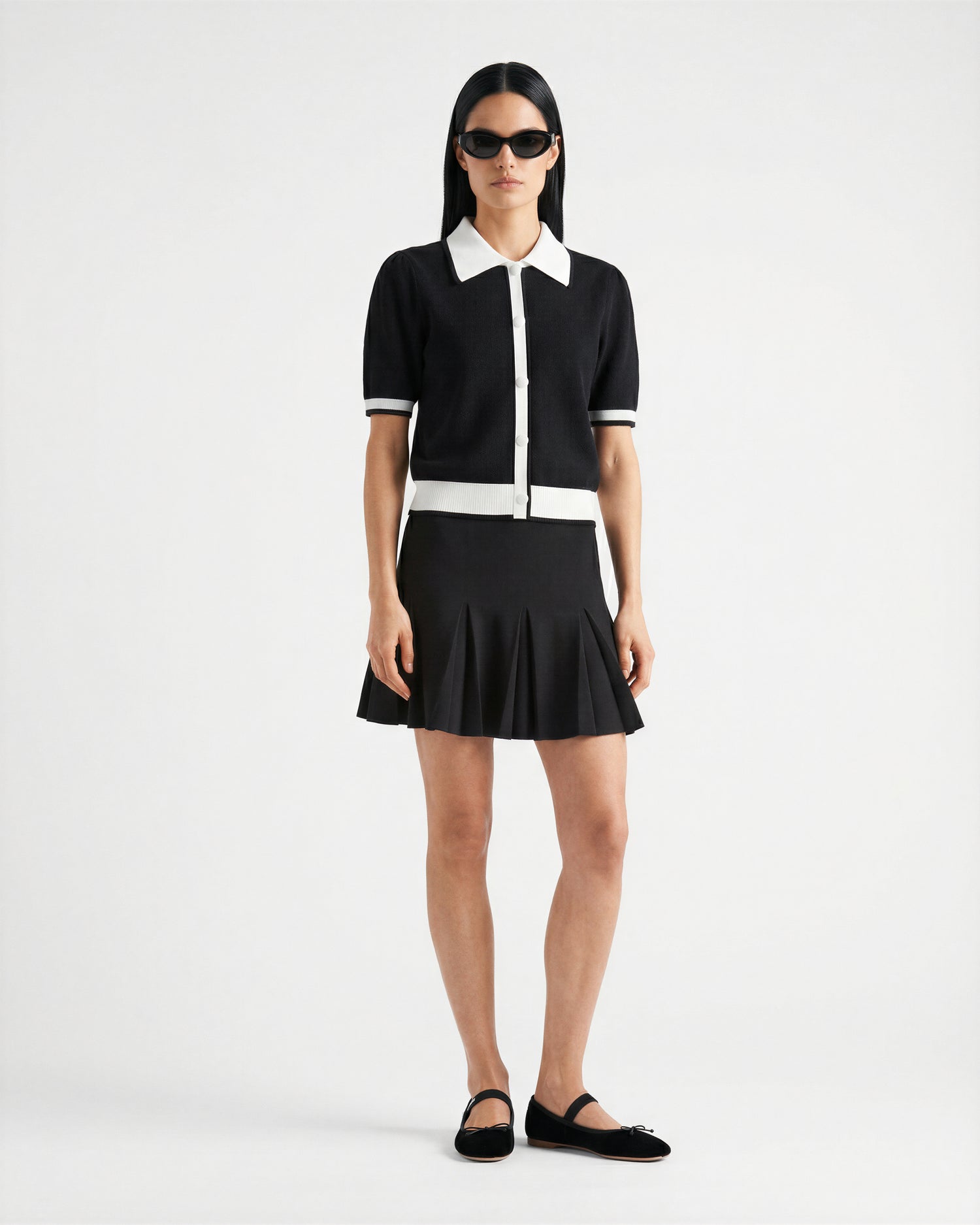 A woman wearing Rudsak’s IRIS - 8526012 Black black pleated mini skirt, a short-sleeve knit sweater with white trim and buttons, black flats, and sunglasses stands against a plain white background.