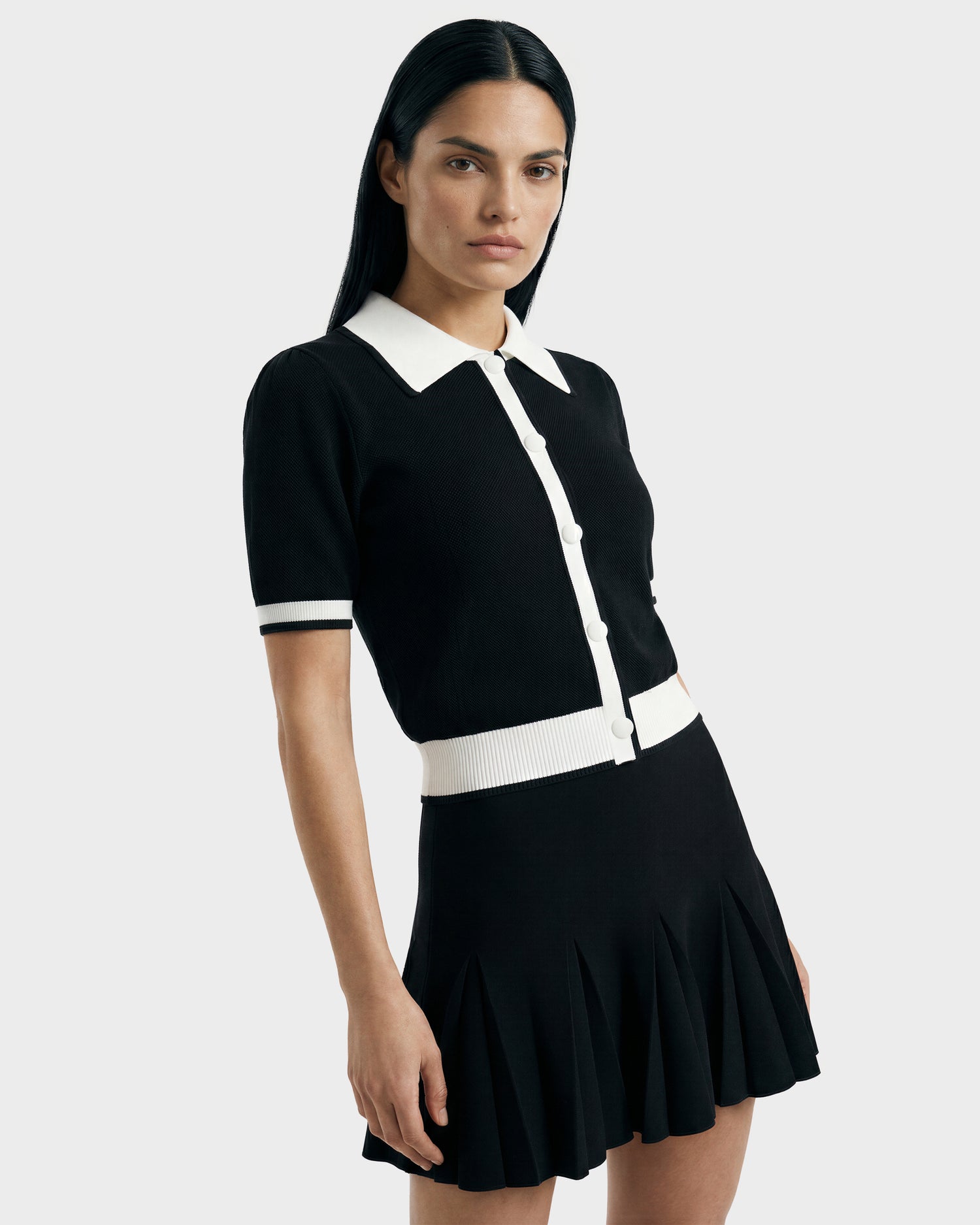 A woman with long dark hair wears the Rudsak IRIS - 8526012 Black knit short-sleeve sweater with contrast collar and white buttons, paired with a black pleated skirt, standing against a plain light background.