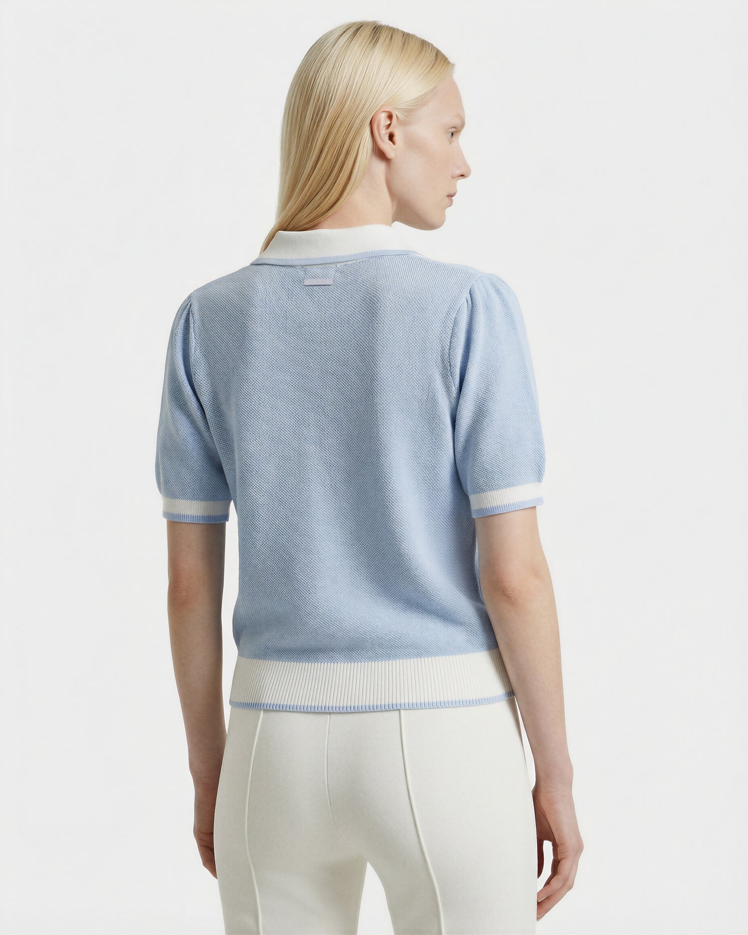 A woman with straight blonde hair is seen from behind, wearing the Rudsak IRIS - 8526012 Powder Blue knit short-sleeve sweater with white trim and high-waisted white pants against a plain white background.