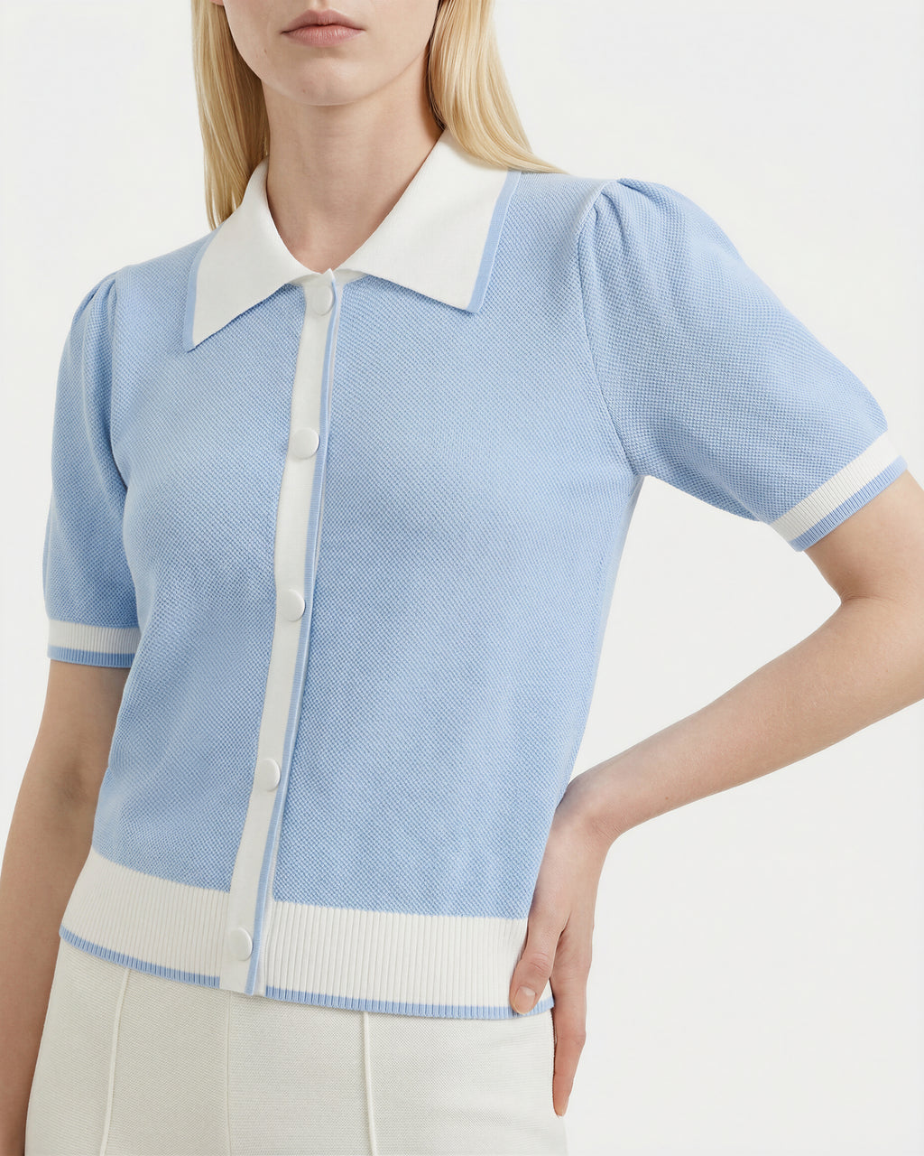 A woman wears the Rudsak IRIS - 8526012 Powder Blue knit short-sleeve sweater, featuring a contrast white collar, buttons, and trim, styled with high-waisted white pants.