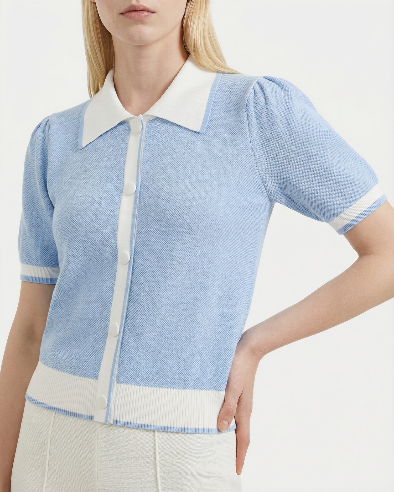 A woman wears the Rudsak IRIS - 8526012 Powder Blue knit short-sleeve sweater, featuring a contrast white collar, buttons, and trim, styled with high-waisted white pants.