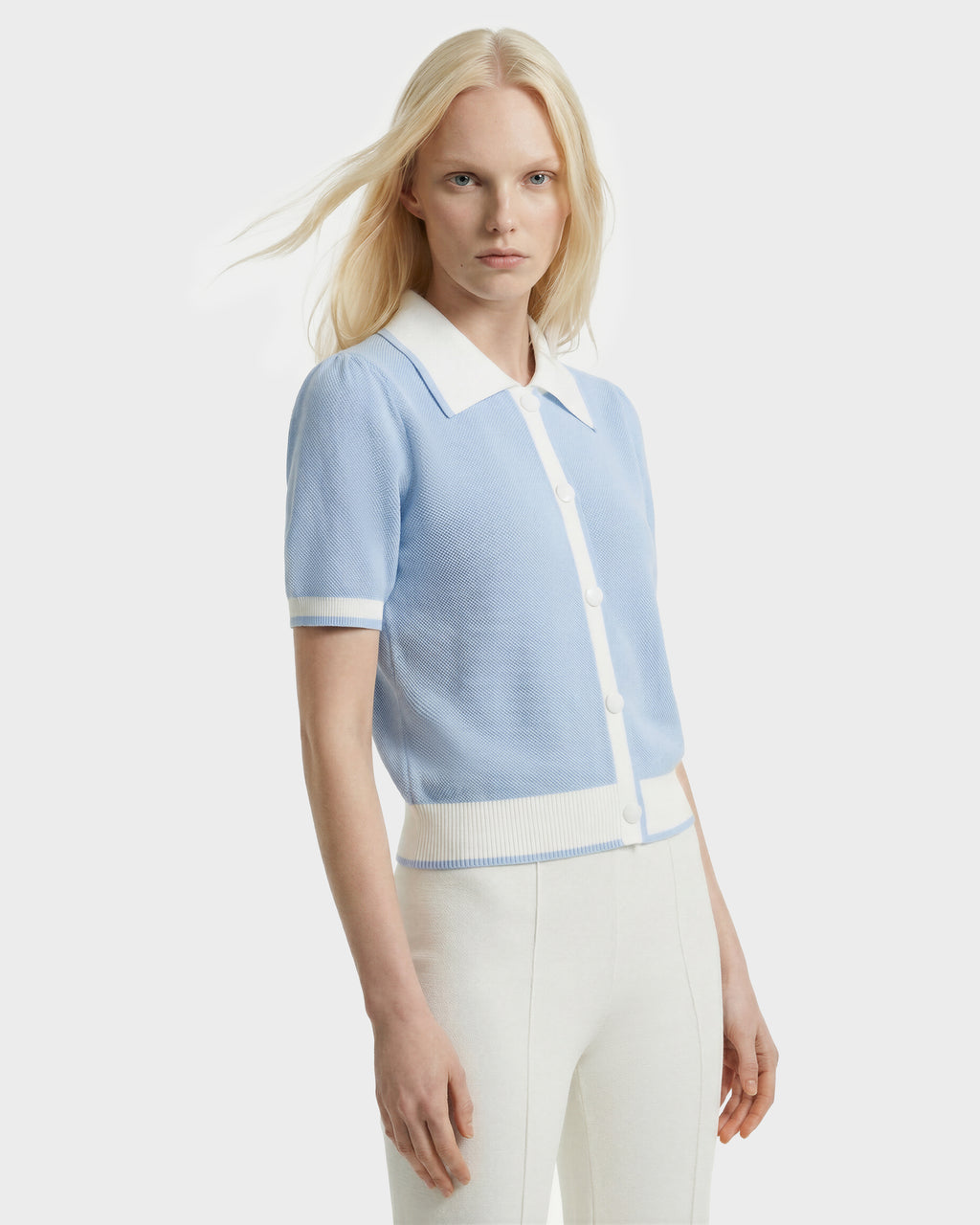 A woman with long blonde hair models the Rudsak IRIS - 8526012 knit short-sleeve sweater in powder blue with white trim and buttons, paired with high-waisted white pants, against a plain white background.