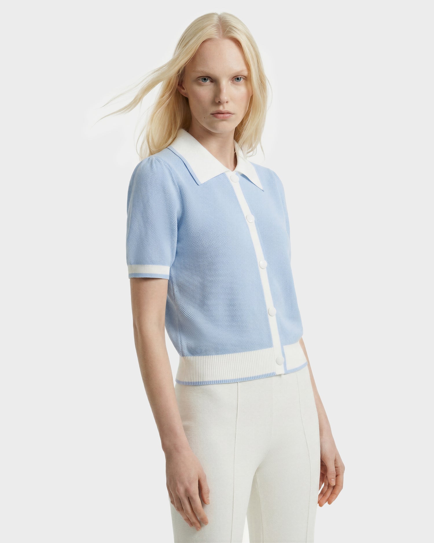 A woman with long blonde hair models the Rudsak IRIS - 8526012 knit short-sleeve sweater in powder blue with white trim and buttons, paired with high-waisted white pants, against a plain white background.
