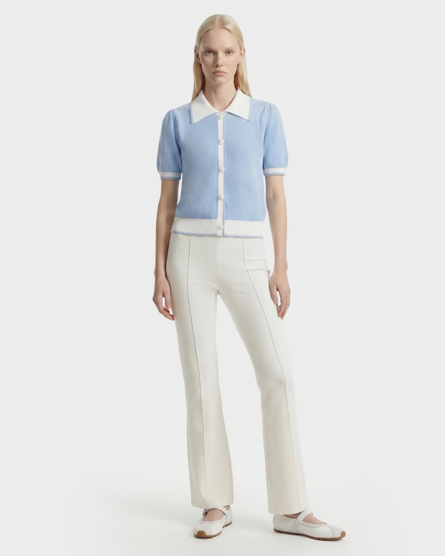 A person with long blonde hair and a neutral expression stands against a white background, wearing the Rudsak IRIS - 8526012 Powder Blue knit short-sleeve sweater featuring white trim, a contrast collar, white fitted pants, and white flats.