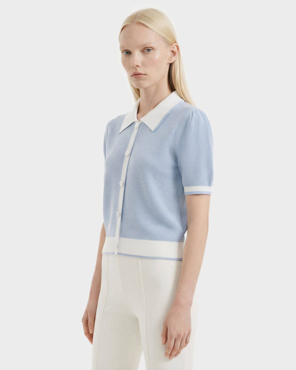 A woman with long blonde hair wears the Rudsak IRIS - 8526012 Powder Blue knit short-sleeve sweater with white trim and buttons, featuring a contrast collar, paired with high-waisted white pants against a plain light background.