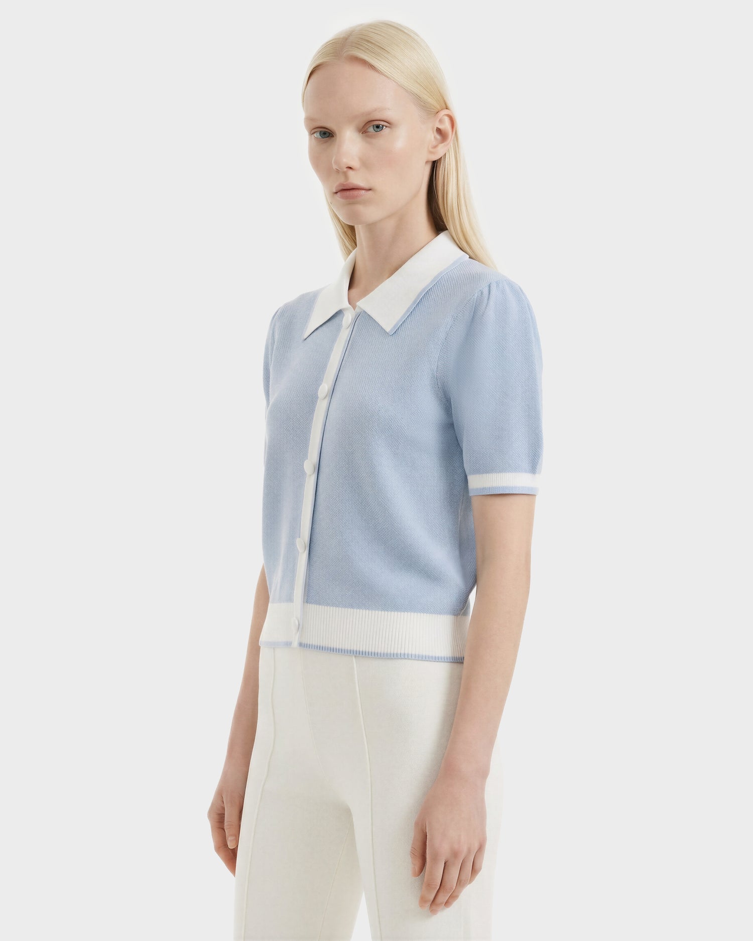 A woman with long blonde hair wears the Rudsak IRIS - 8526012 Powder Blue knit short-sleeve sweater with white trim and buttons, featuring a contrast collar, paired with high-waisted white pants against a plain light background.