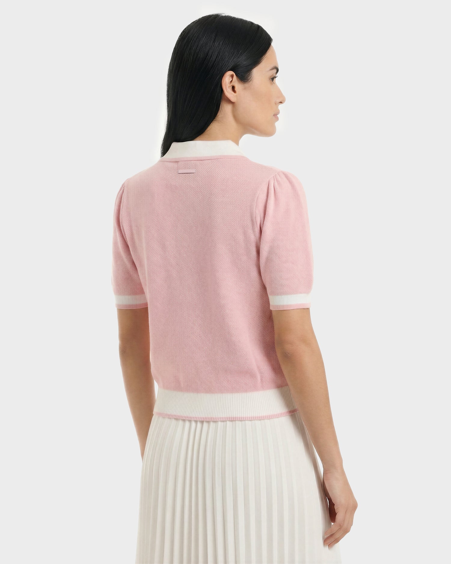 A woman with straight dark hair is shown from the back, wearing the Rudsak IRIS - 8526012 Pink light pink knit short-sleeve sweater with white trim and a white pleated skirt, standing against a plain light background.