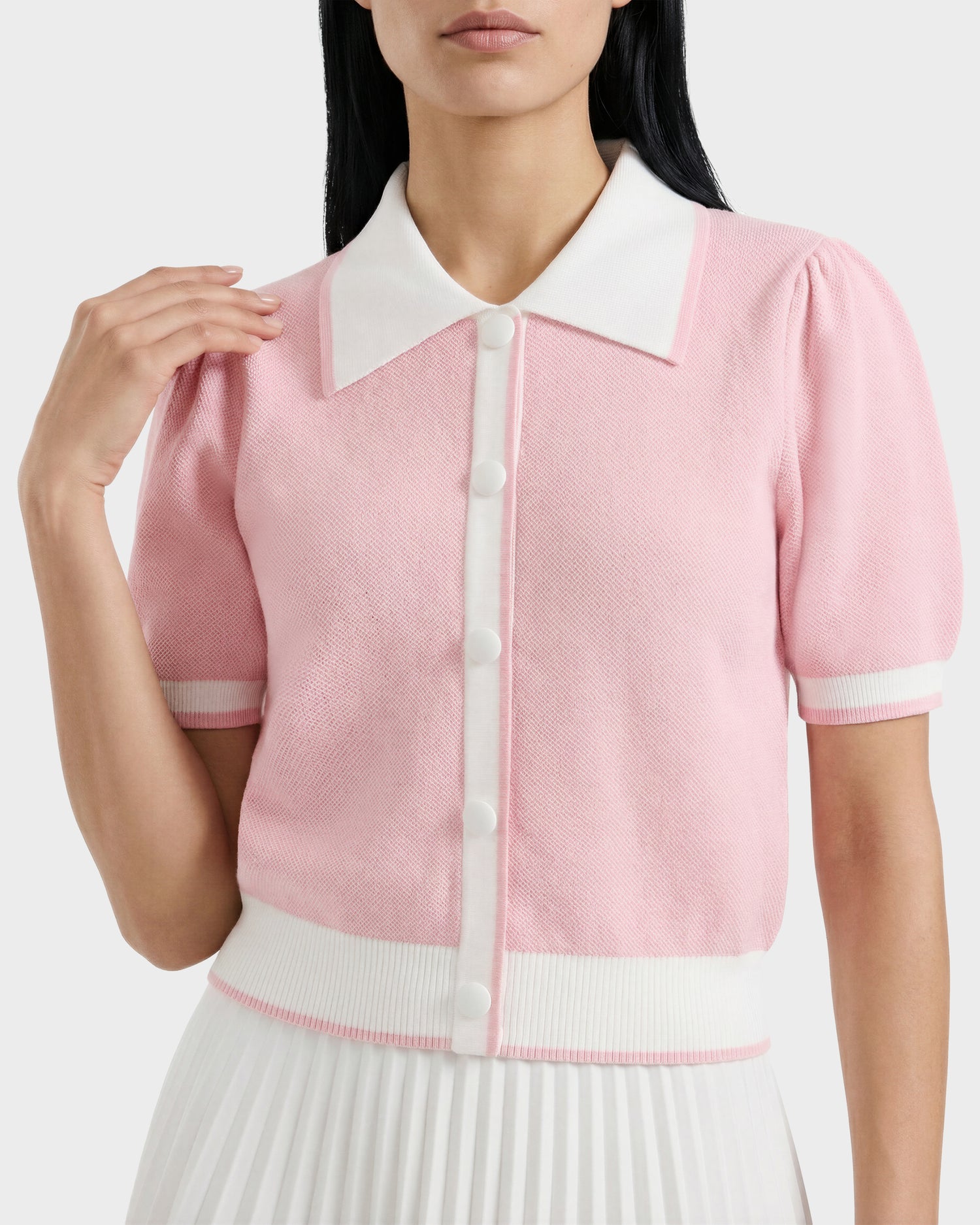 A woman wears the Rudsak IRIS - 8526012 Pink knit short-sleeve sweater with white buttons, a contrast collar, and white trim, paired with a white pleated skirt. Her face is partially out of frame.