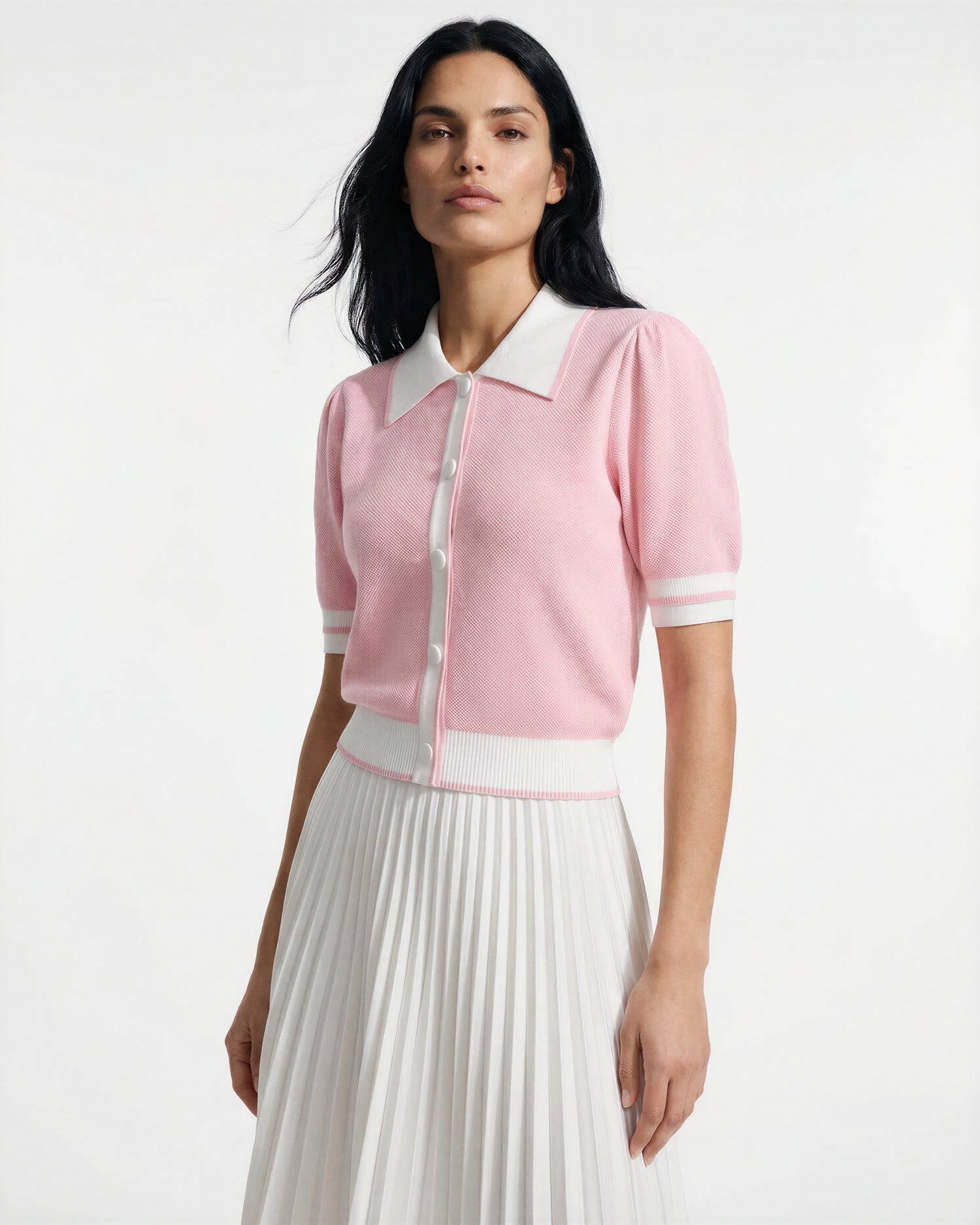 A woman stands against a plain white background, wearing the Rudsak IRIS - 8526012 Pink knit short-sleeve sweater with contrast collar, white trim, and buttons, paired with a white pleated skirt.