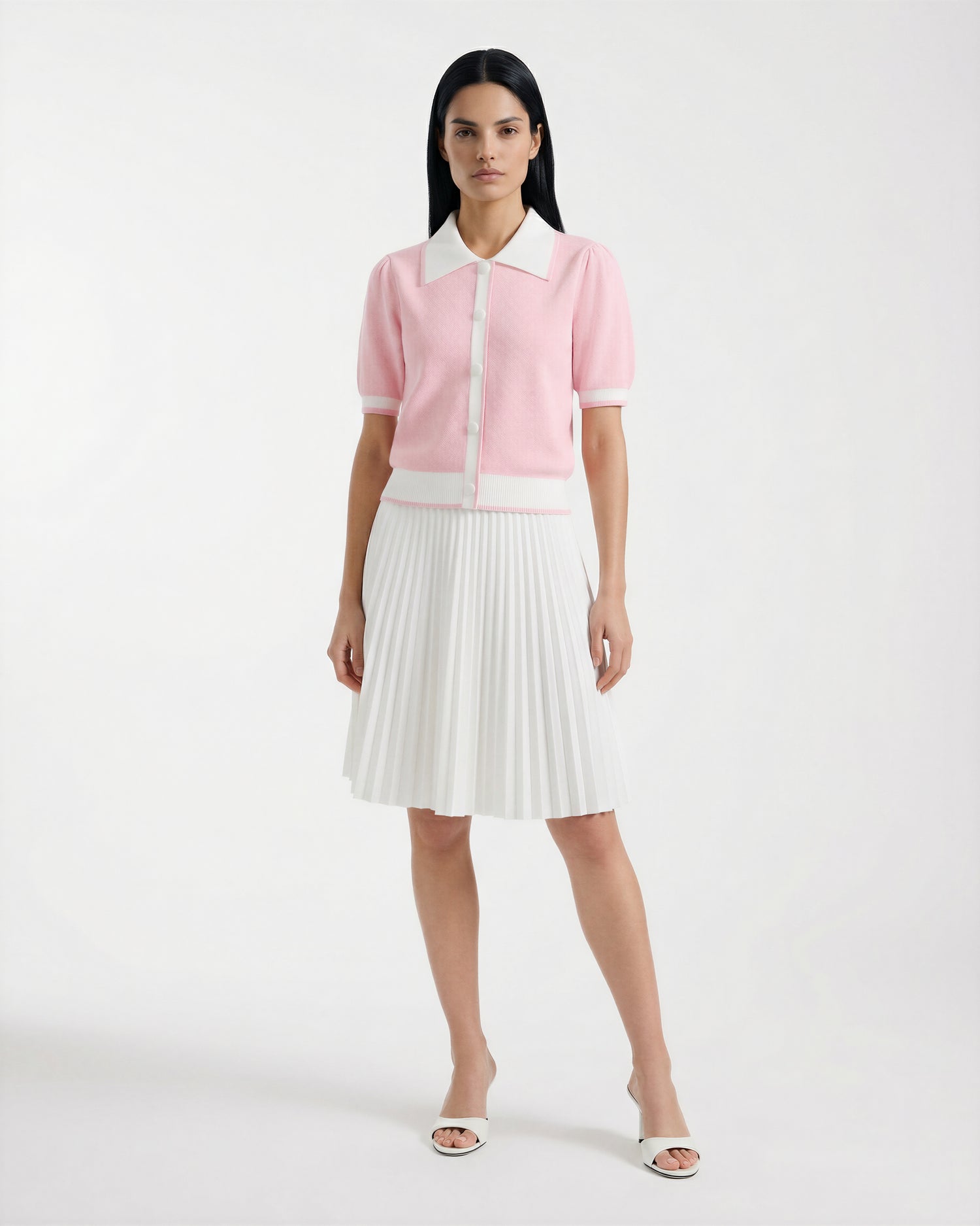 A woman wears the Rudsak IRIS - 8526012 Pink knit short-sleeve sweater with a contrast collar, styled with a white pleated skirt and white heeled sandals, standing against a plain white background.