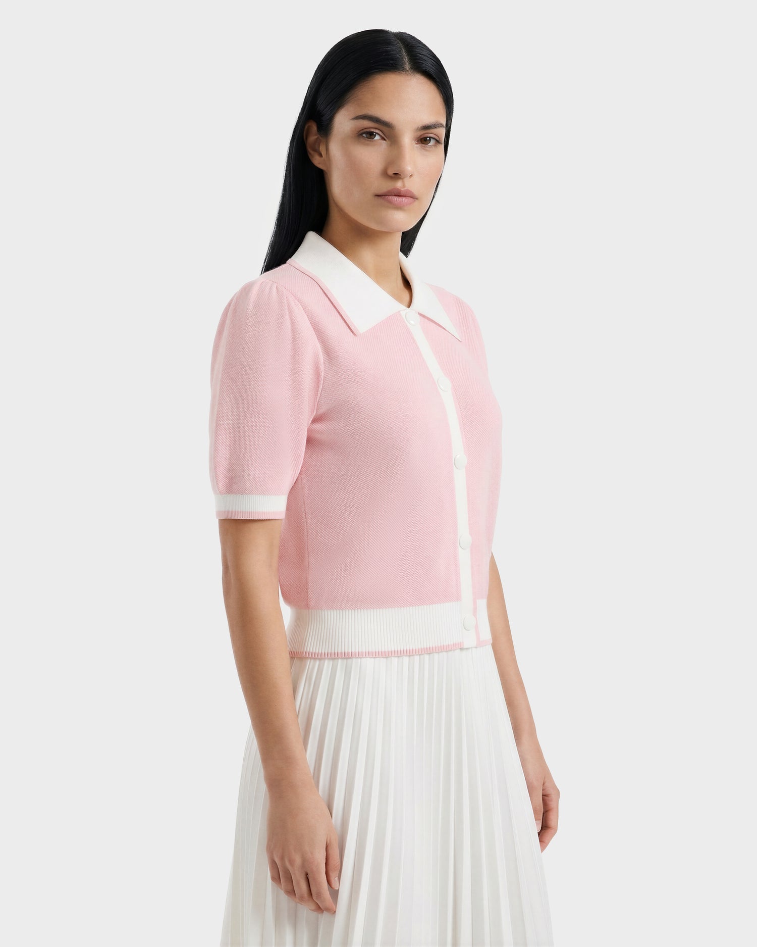 A woman with long dark hair wears the IRIS - 8526012 Pink short-sleeve sweater by Rudsak, featuring a contrast collar, white buttons, and white trim, paired with a white pleated skirt against a plain light background.