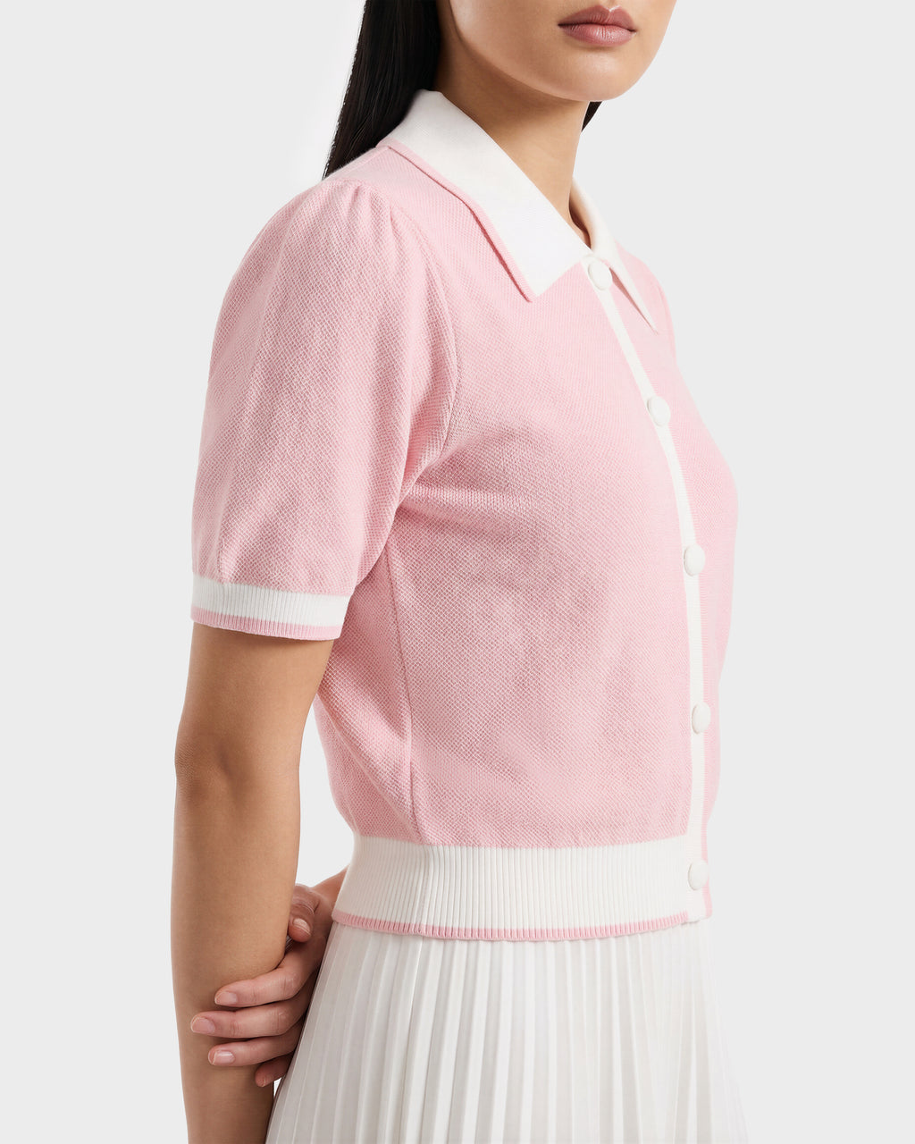 A woman wears the Rudsak IRIS - 8526012 light pink knit short-sleeve sweater, featuring a contrast collar and white trim, paired with a white pleated skirt. She stands in profile with her hands clasped behind her back.