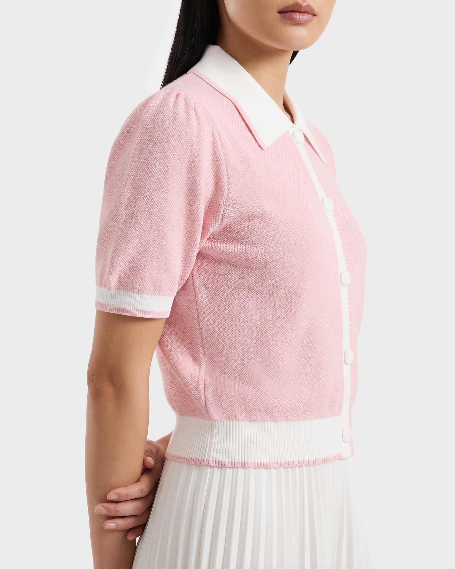 A woman wears the Rudsak IRIS - 8526012 light pink knit short-sleeve sweater, featuring a contrast collar and white trim, paired with a white pleated skirt. She stands in profile with her hands clasped behind her back.