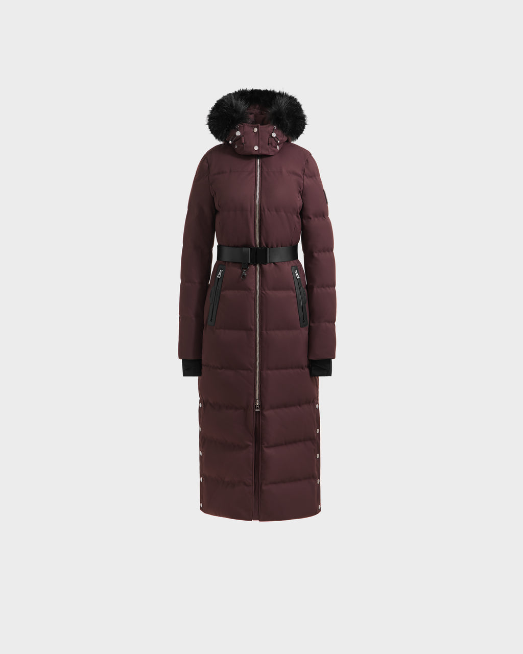 The Rudsak IZABELLA H - 8125644 Bordeaux is a long maroon women’s puffer coat with a black faux fur-trimmed hood, front zipper, black waist belt, and zippered pockets, shown against a plain white background.