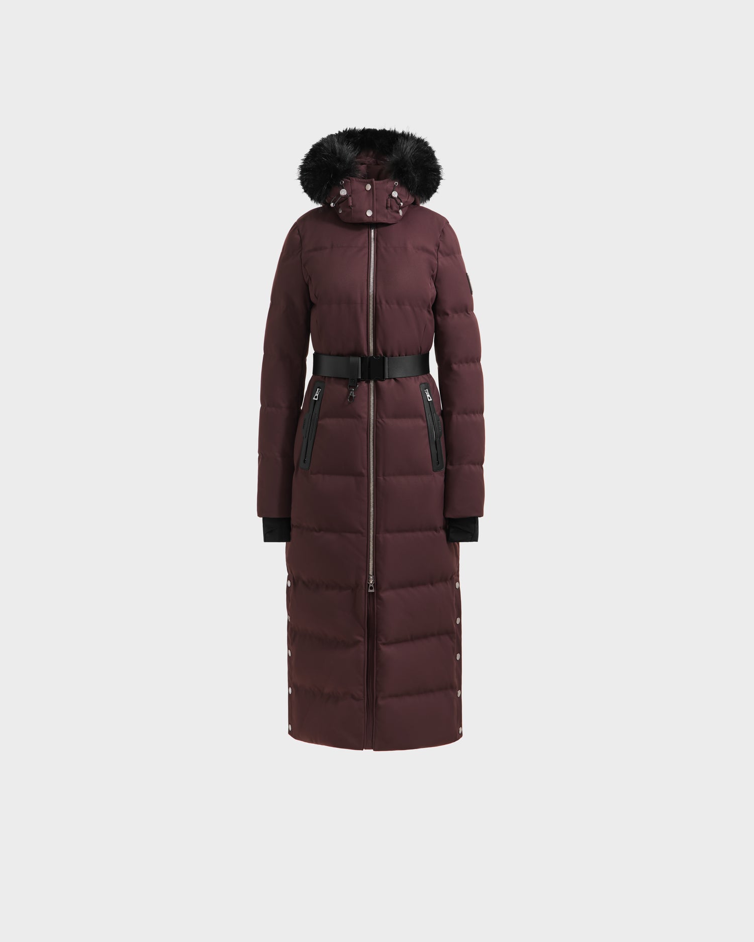 The Rudsak IZABELLA H - 8125644 Bordeaux is a long maroon women’s puffer coat with a black faux fur-trimmed hood, front zipper, black waist belt, and zippered pockets, shown against a plain white background.