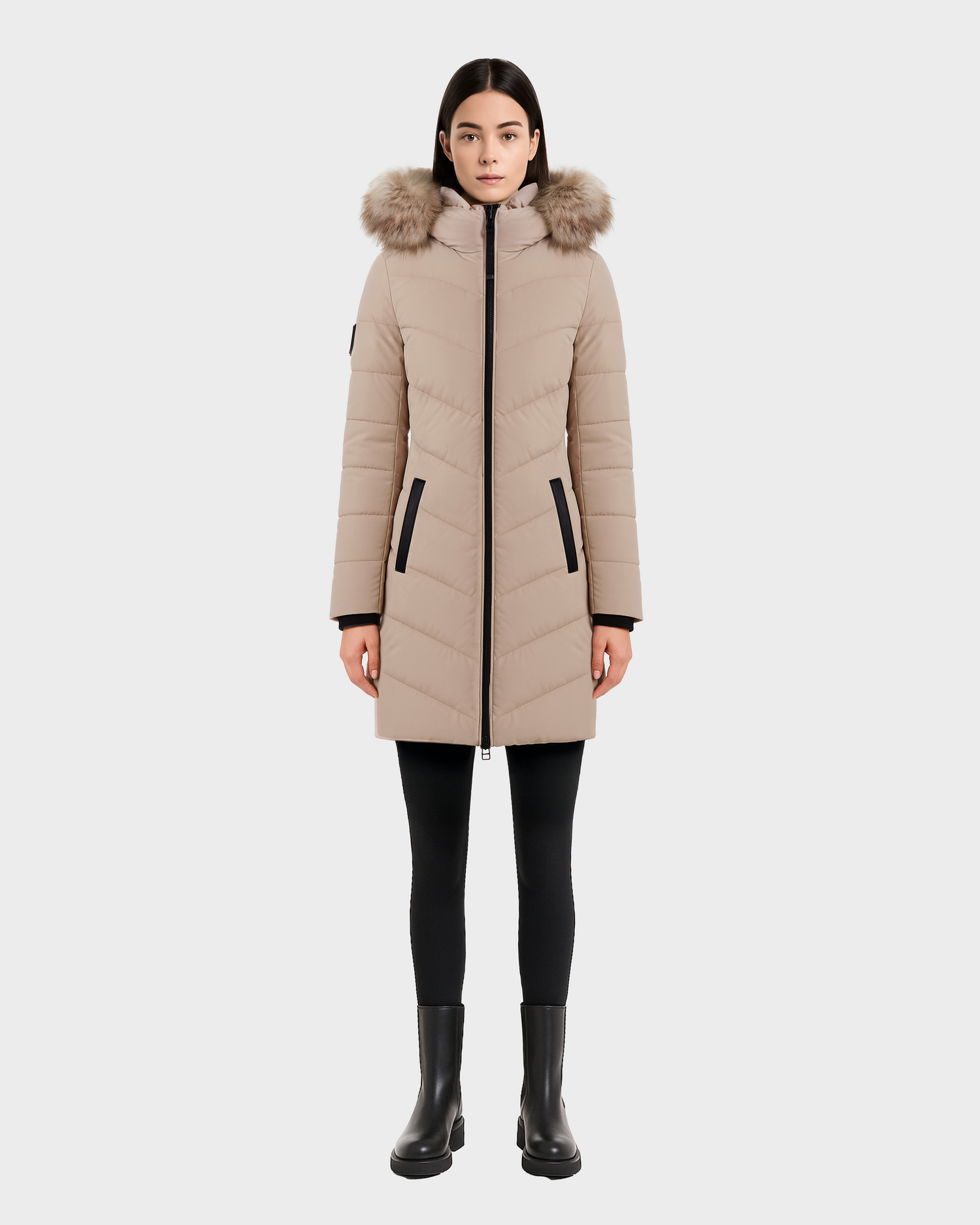 A woman faces forward in the Rudsak LAUREN - 4124520 Taupe knee-length quilted down coat with a faux fur hood, paired with black leggings and ankle boots, set against a plain light background.