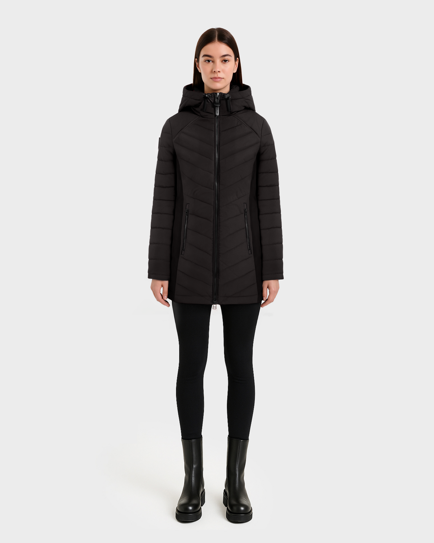 A woman stands facing forward, wearing the Rudsak CORDELIA - 4124519 Black down puffer jacket with a hood, paired with black leggings and boots against a plain light background.