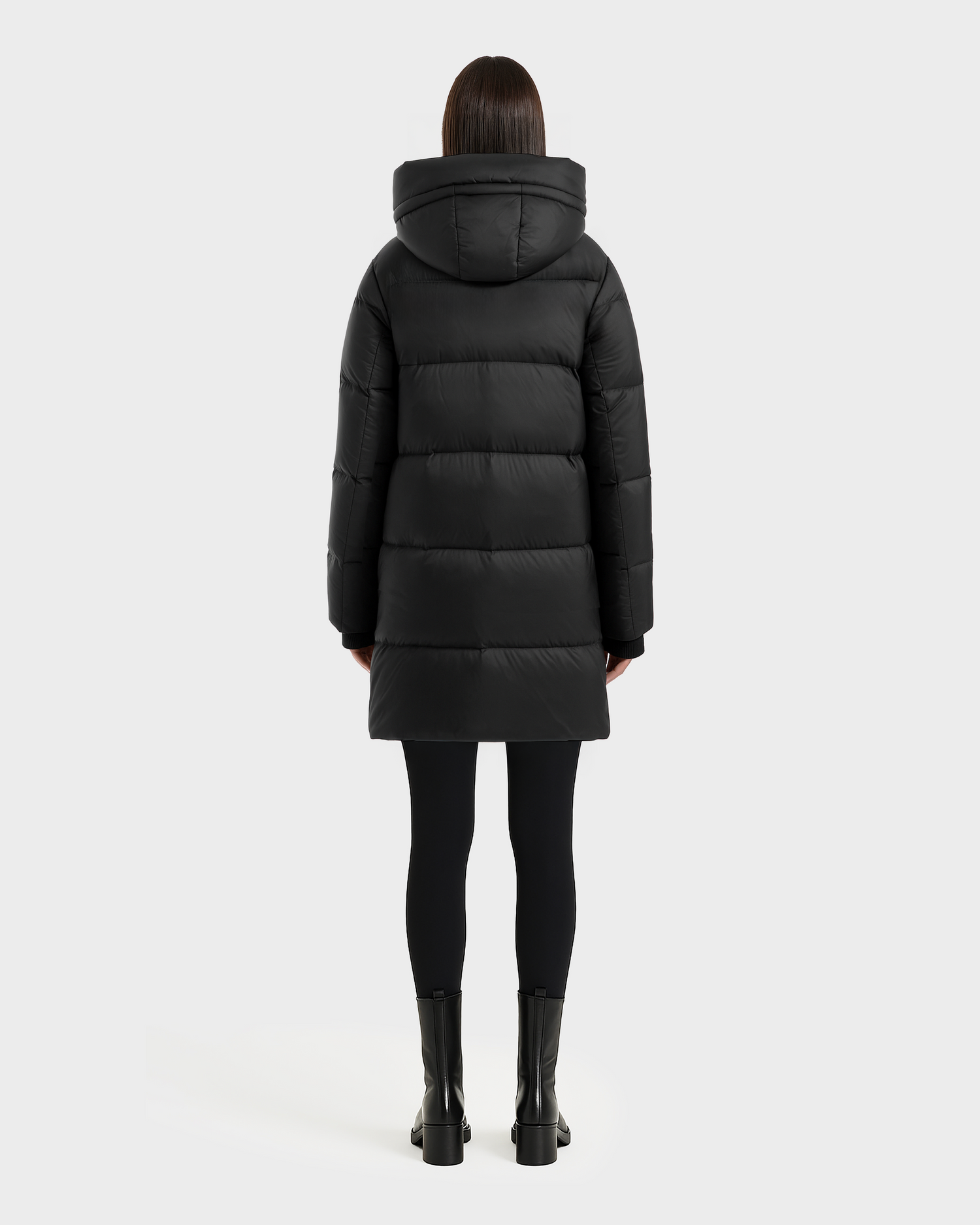 A person with straight dark hair, seen from behind, is wearing the Rudsak OLIVIA - 4123509 Black puffer coat with a hood, along with black leggings and ankle boots, standing against a plain light gray background.