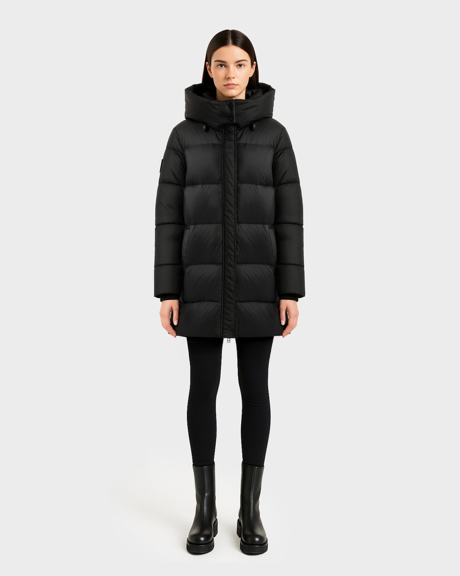 A woman faces forward against a plain light gray background, wearing the Rudsak OLIVIA - 4123509 Black long puffer jacket with high collar and hood, paired with black leggings and ankle boots.