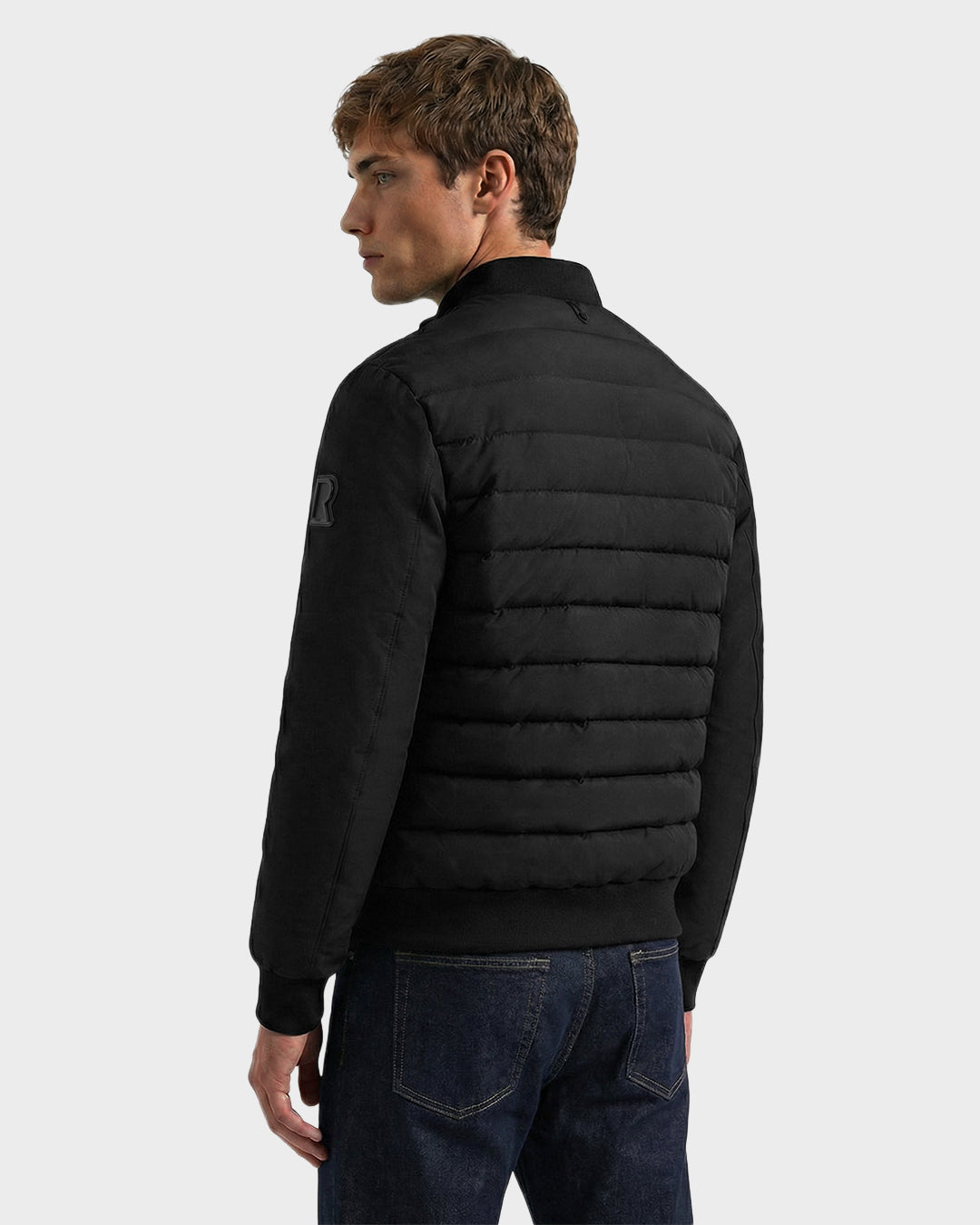 A young man with short brown hair stands facing away, wearing the Rudsak JIMMY - 6125723 Black men's quilted down jacket and dark blue jeans against a plain light gray background.