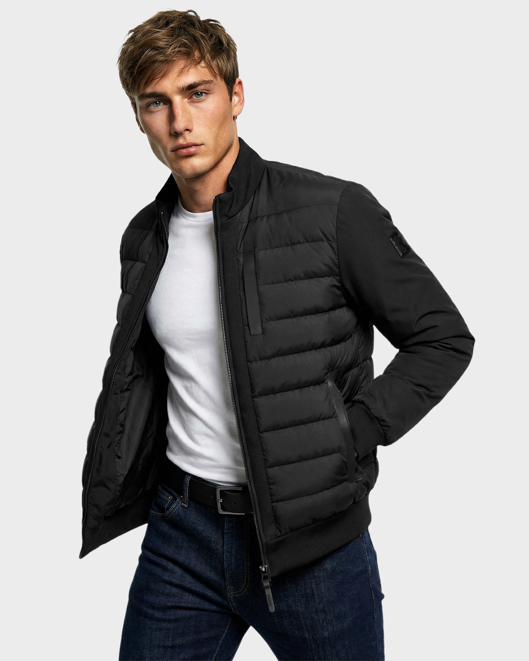 A young man with short brown hair models the Rudsak JIMMY - 6125723 Black men's down jacket—water resistant and stylish—over a white t-shirt and dark jeans, standing with hands in pockets against a plain light gray background.