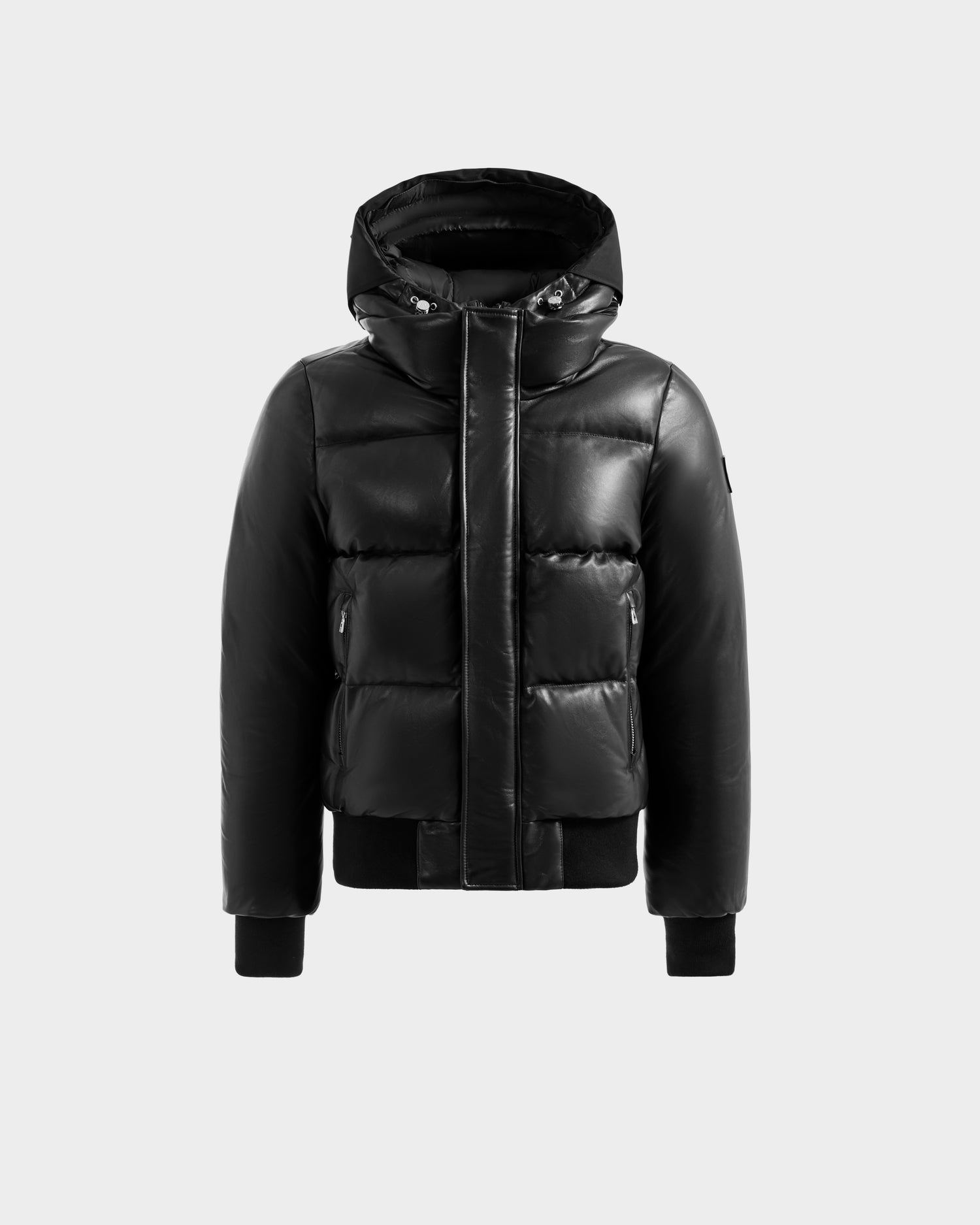 The Rudsak JORDY X - 6125694 Black is a black, hooded quilted bomber puffer jacket with a shiny finish, front zipper under a snap-button flap, zippered side pockets, and ribbed cuffs and hem, shown on a light gray background.