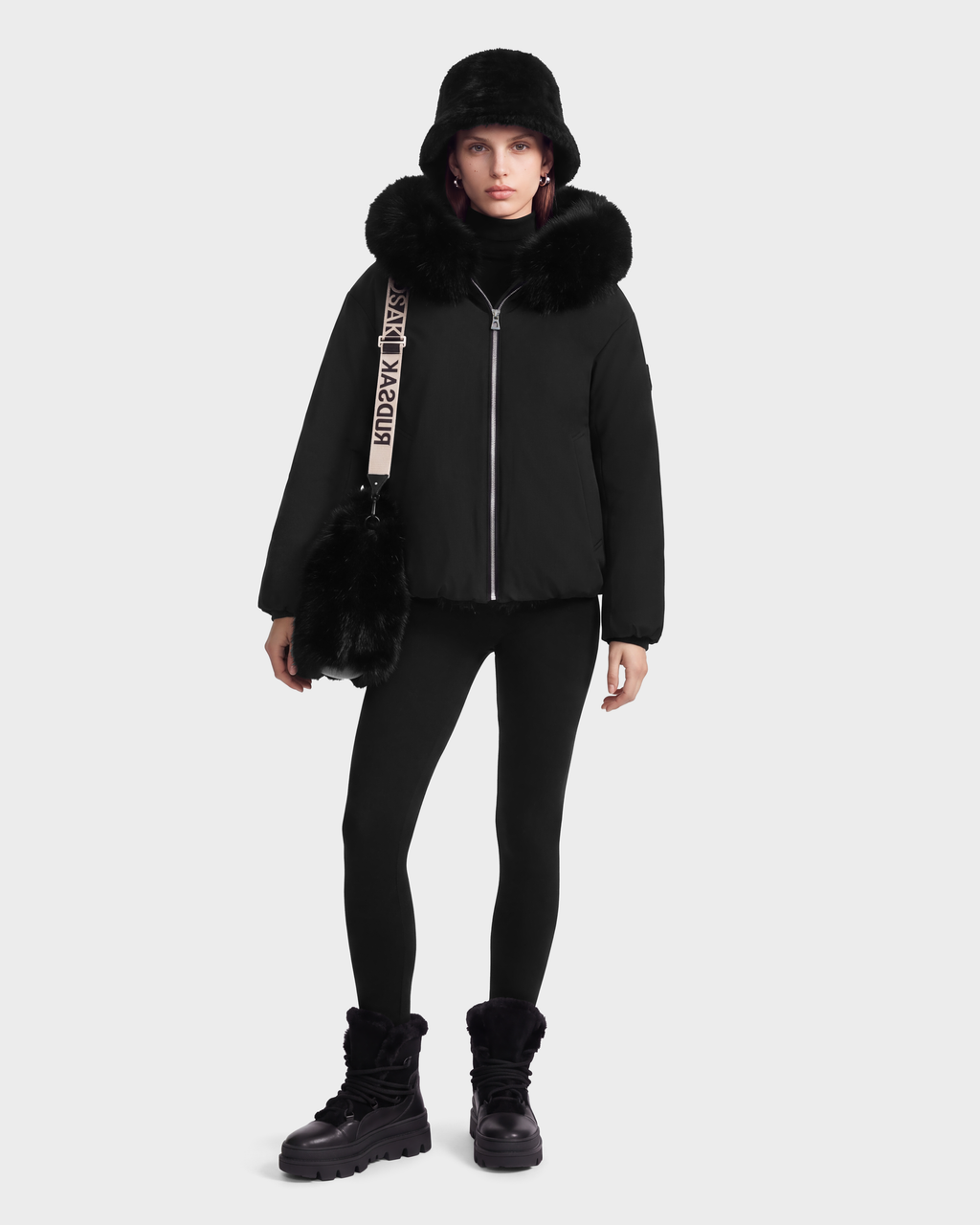 A person wearing a KATHLEEN - 8126764 Black puffer jacket with a fur-trimmed hood, a bucket hat, black leggings, and black boots, carrying a black bag with a branded shoulder strap.