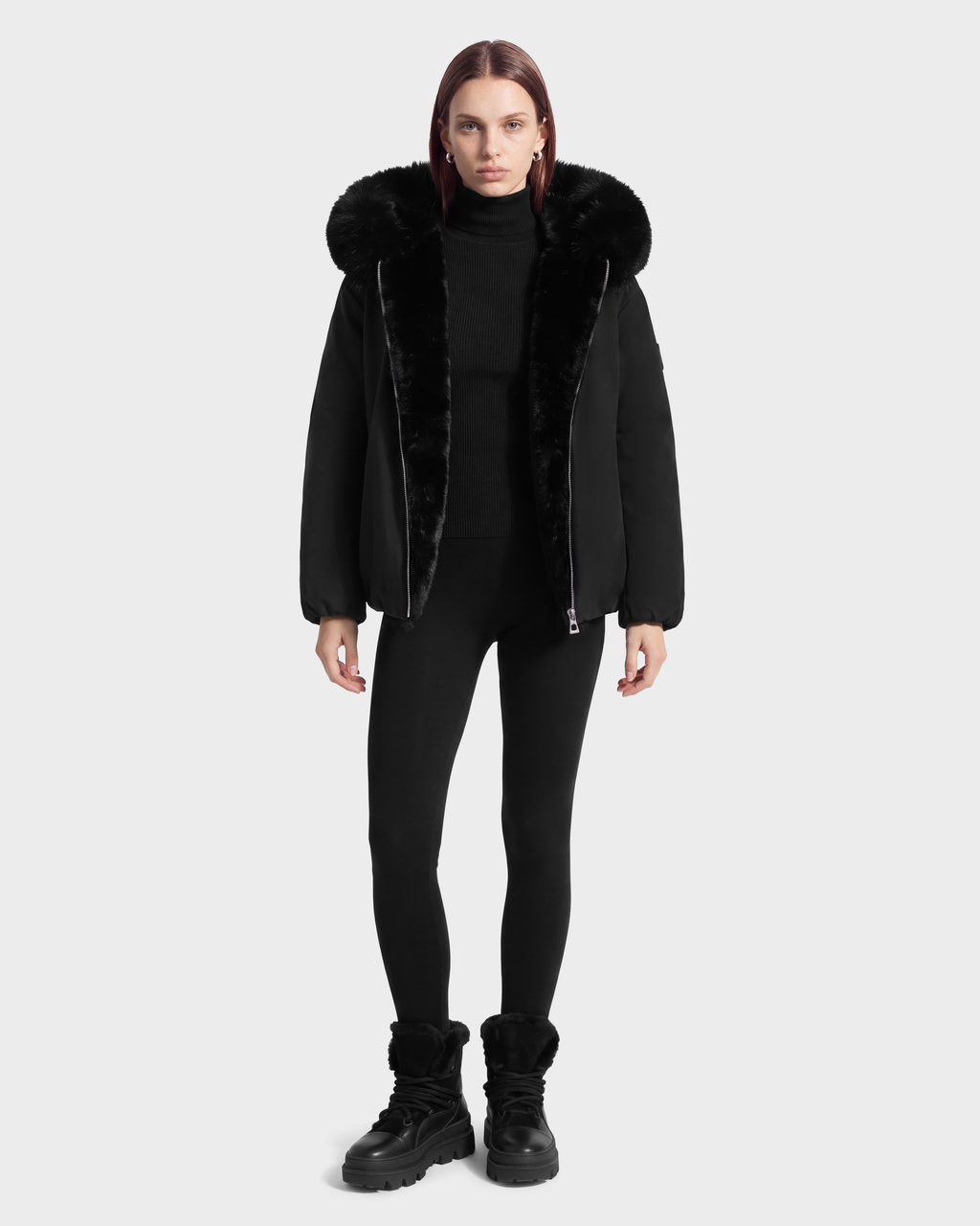 A model wearing a KATHLEEN - 8126764 Black winter jacket with a fur-lined hood, a black turtleneck, black leggings, and black winter boots.
