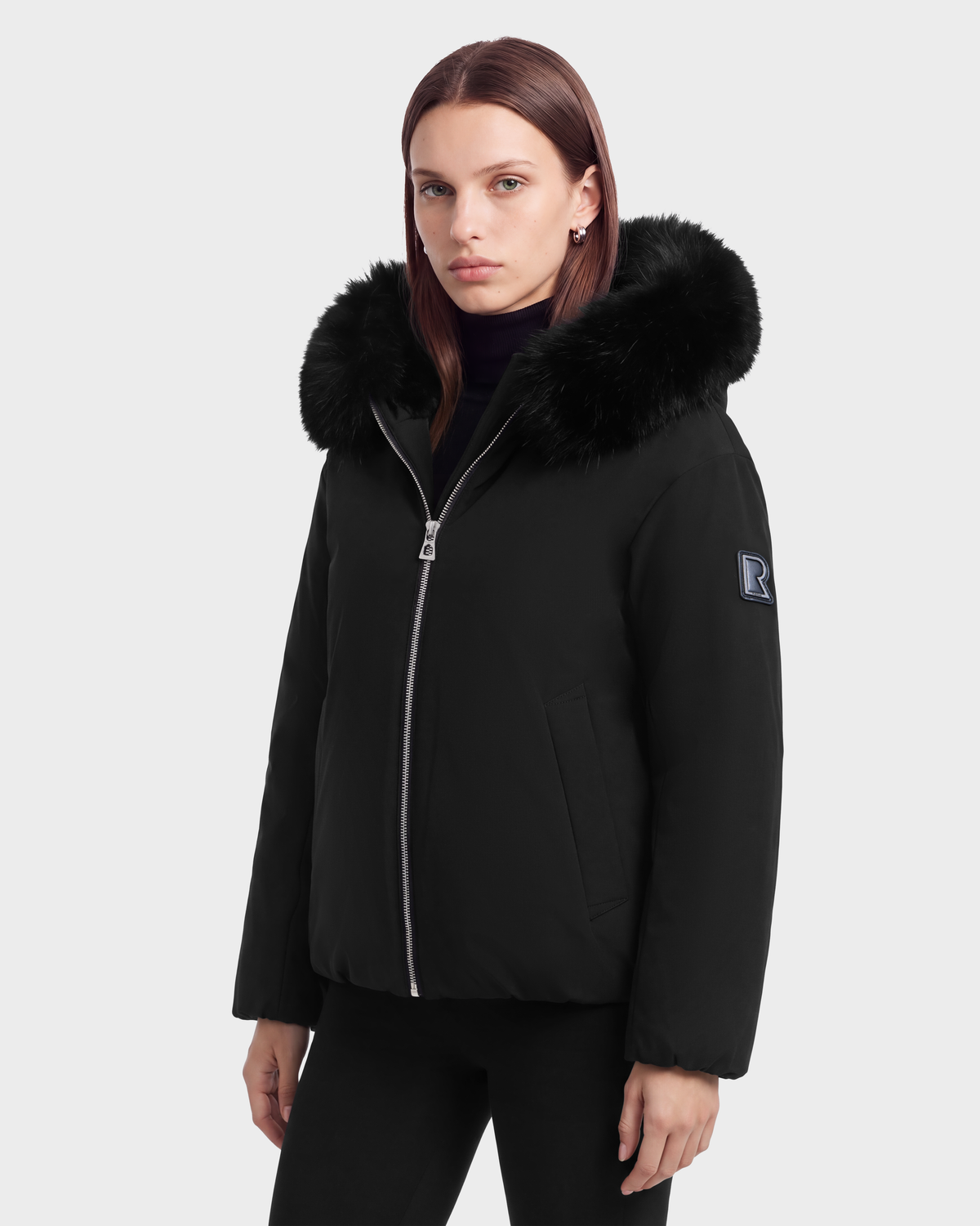 A person wearing a black Rudsak KATHLEEN - 8126764 winter jacket with a fur-trimmed hood and a square logo patch on the sleeve.