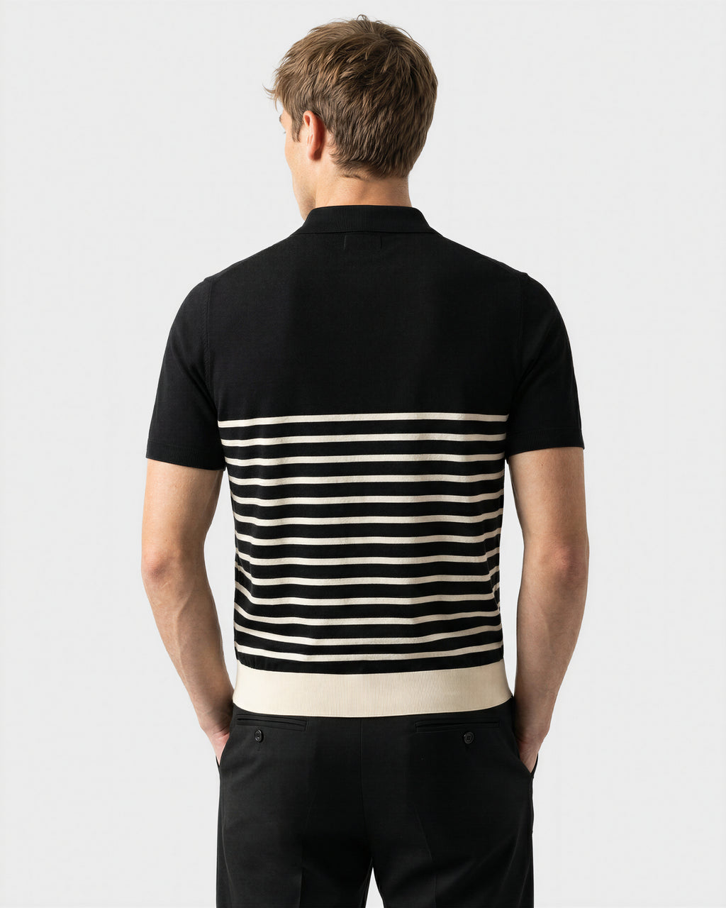 A man with short light brown hair is seen from behind, wearing the Rudsak KRIS - 6526005 Black fine-gauge knit polo with a graphic edge and contrast stripe across the lower back, paired with matching black pants.