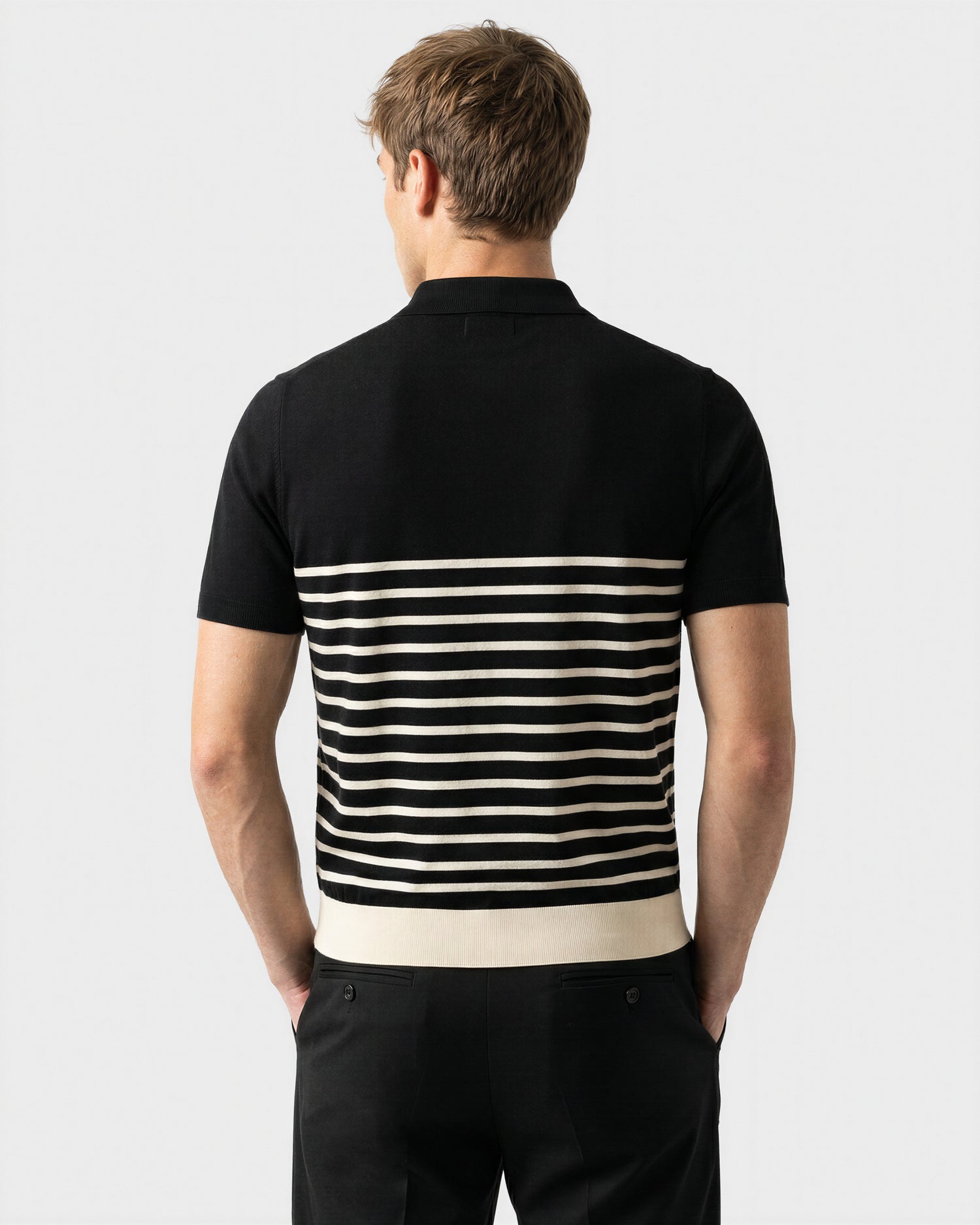A man with short light brown hair is seen from behind, wearing the Rudsak KRIS - 6526005 Black fine-gauge knit polo with a graphic edge and contrast stripe across the lower back, paired with matching black pants.