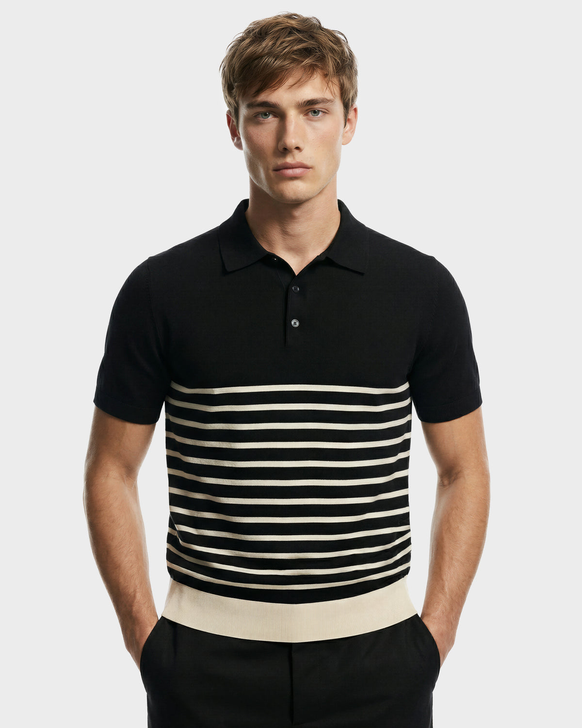 A young man wears the Rudsak KRIS - 6526005 Black contrast stripe polo, featuring horizontal white chest stripes and a solid white hem band, standing with his hands in his pockets against a plain background.