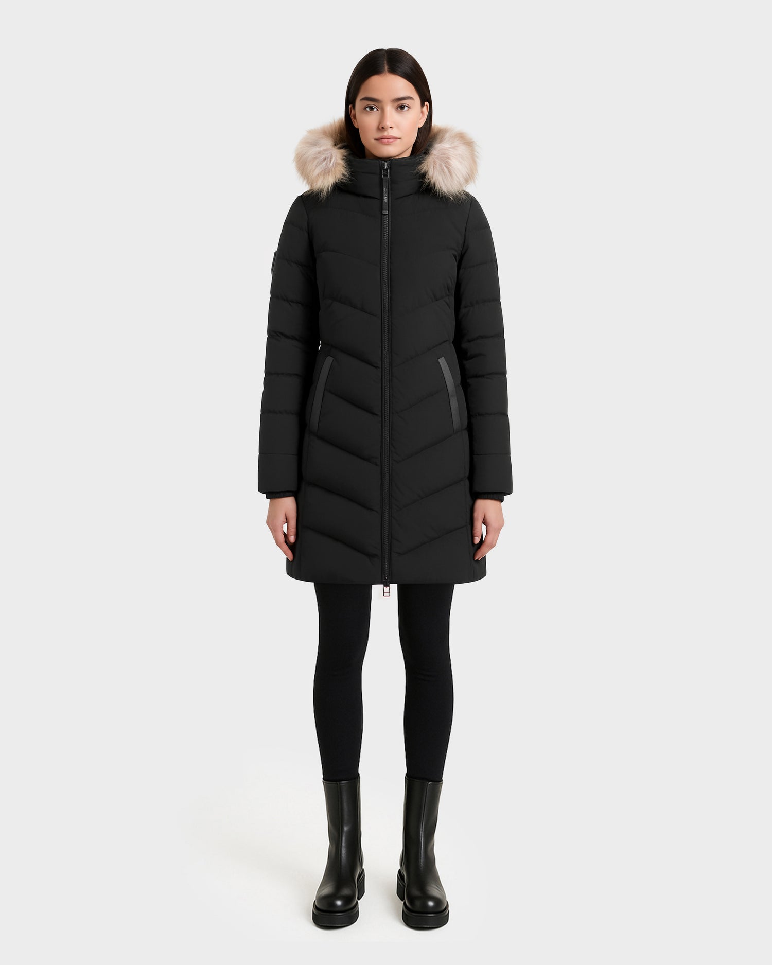 A woman faces forward against a plain light gray background, wearing the Rudsak LAUREN - 4124520 Black long down coat with a faux fur hood, paired with black leggings and black ankle boots.
