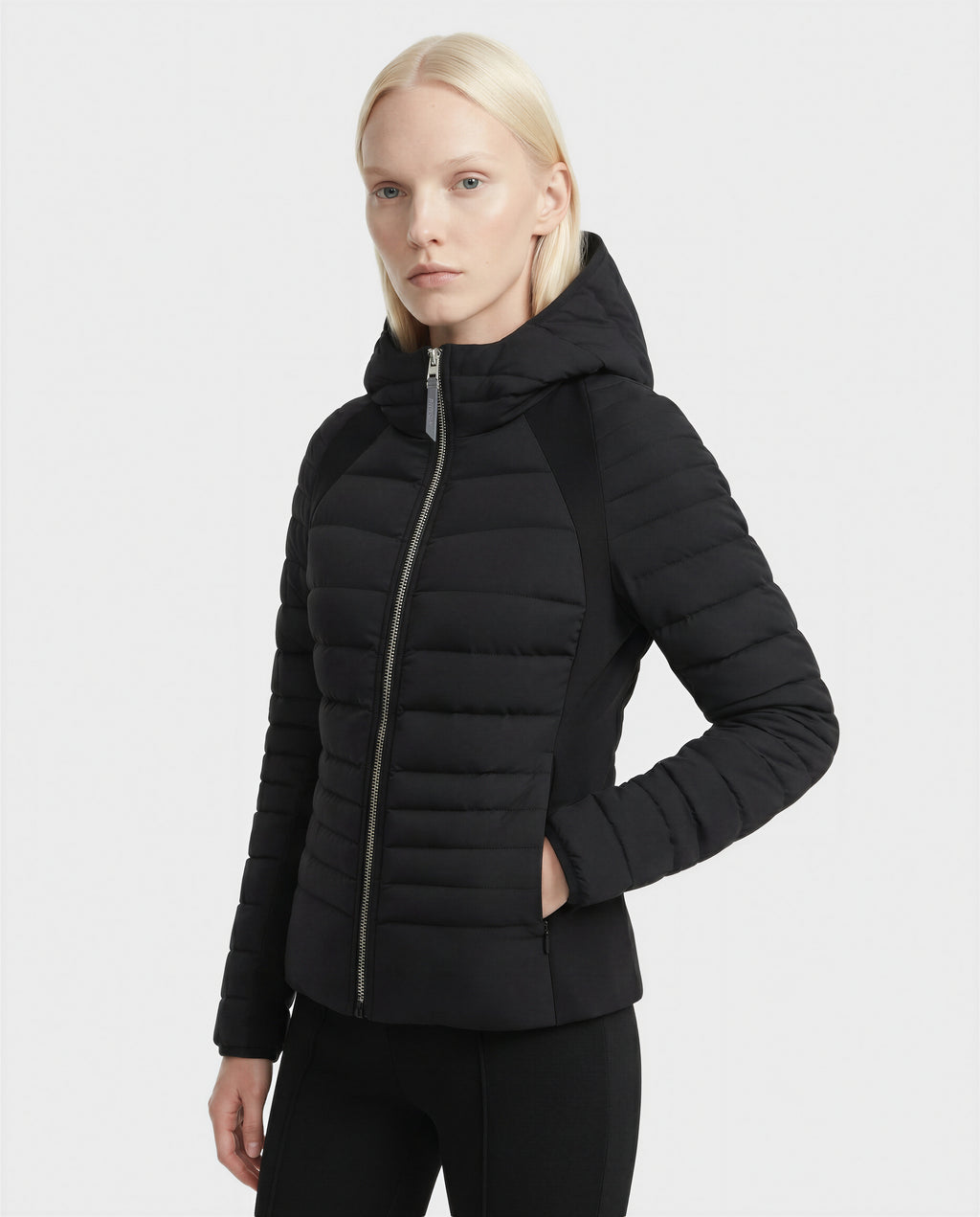 A person with straight, light blonde hair wears the Rudsak LENA - L124005 Black women's fitted light down jacket with hood and neoprene inserts, standing against a plain white background.