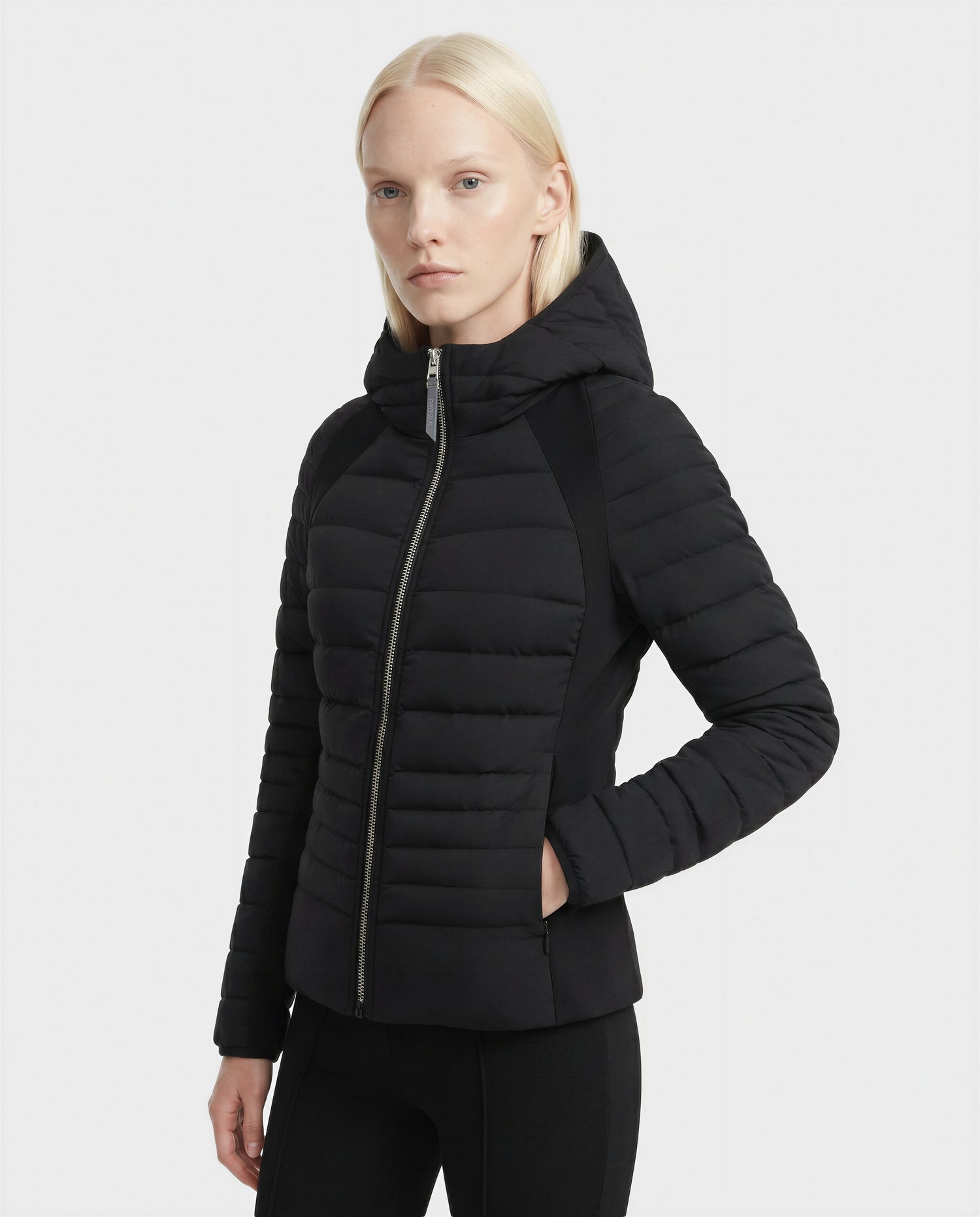 A person with straight, light blonde hair wears the Rudsak LENA - L124005 Black women's fitted light down jacket with hood and neoprene inserts, standing against a plain white background.