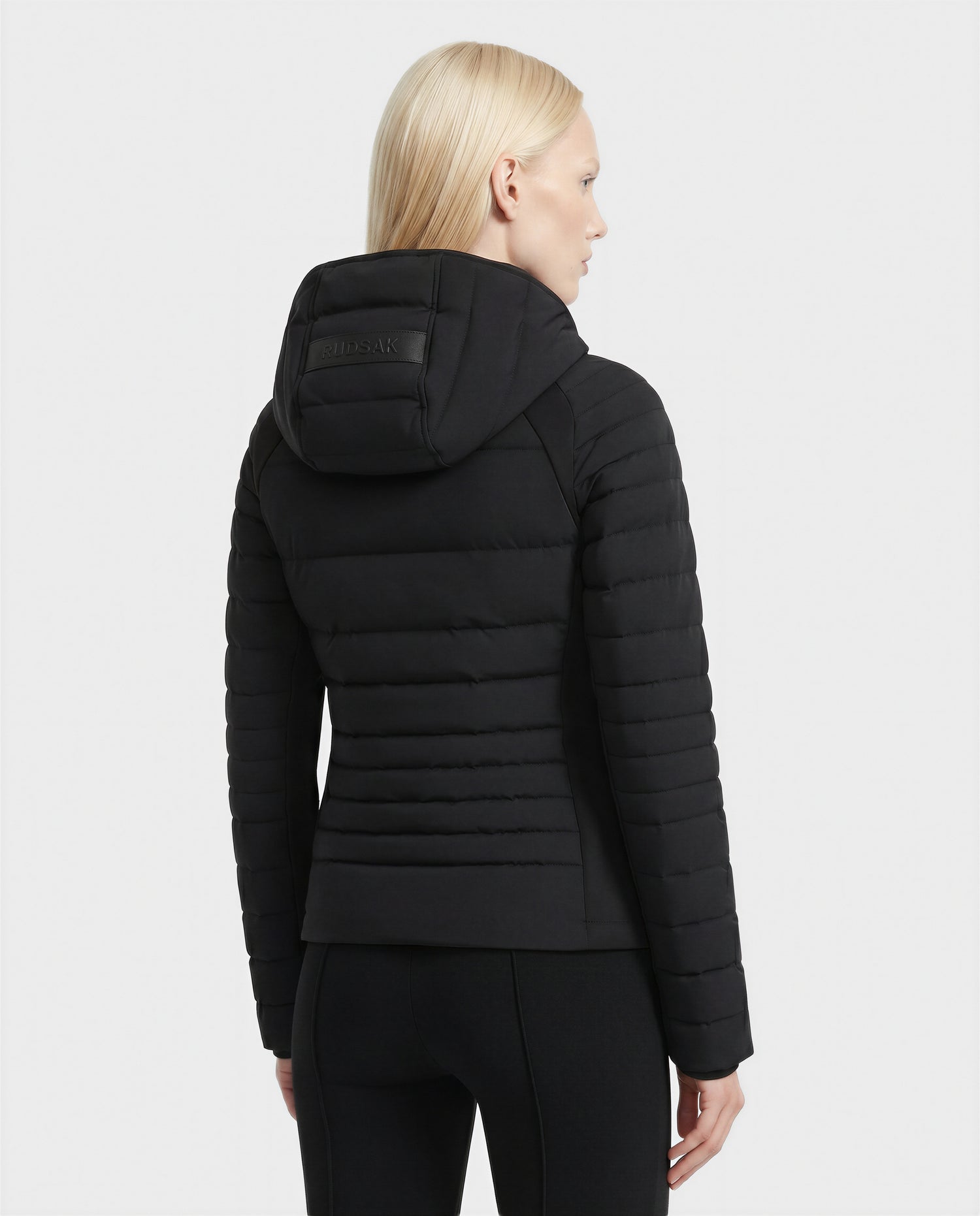 A person with straight, light blonde hair is seen from the back wearing Rudsak's LENA - L124005 Black fitted women’s down jacket with a hood and black pants, set against a plain white background.